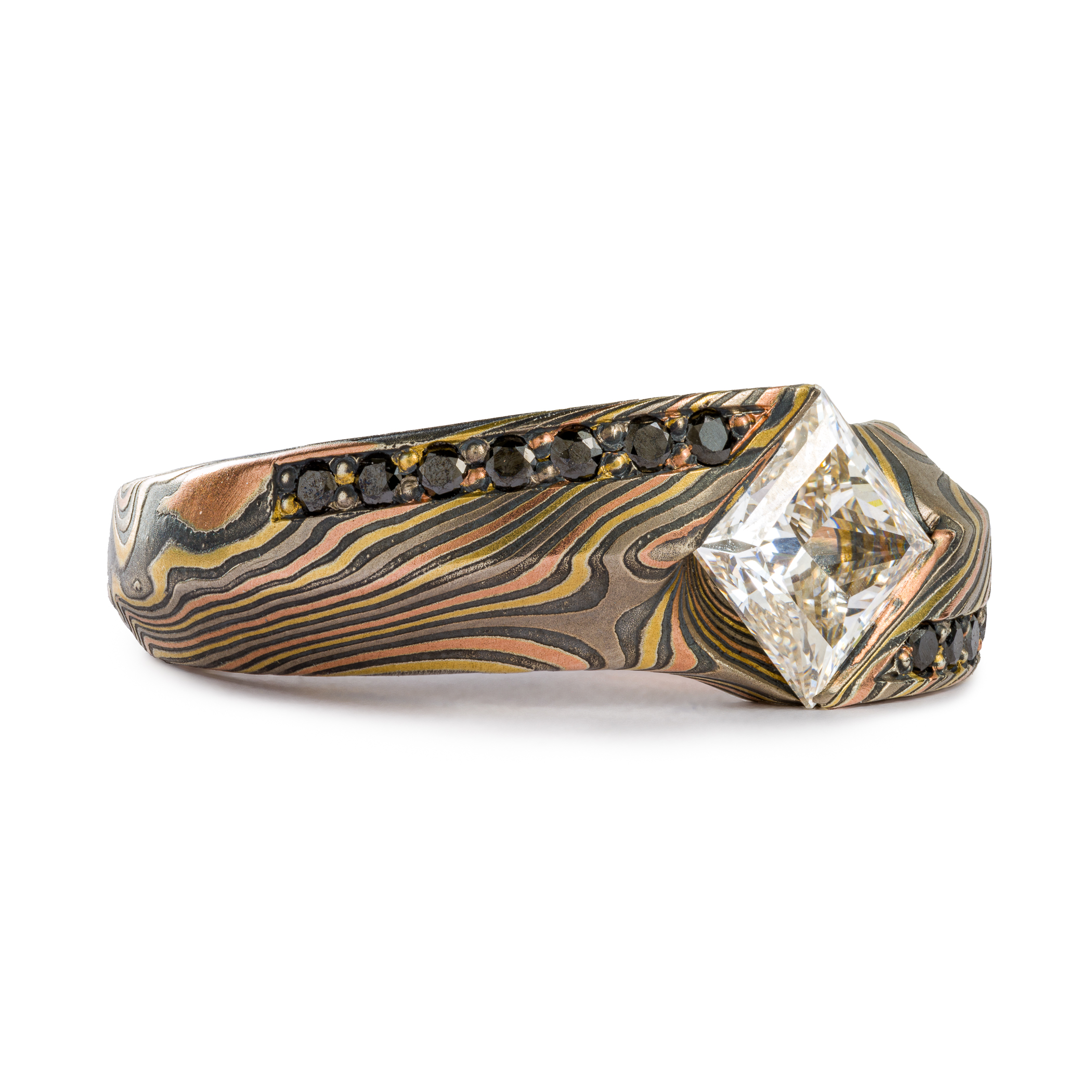 This is a custom made Mokume Gane ring, made by arn krebs, featuring a unique carved angled profile and a modified cathedral style setting holding the large white center diamond. The diamond is square and set diagonally to the band, and is flanked on each side by seven small bead set black diamonds running parallel to the band. It's made in our Twist pattern and an upgraded 18kt version of our Firestorm palette, the metals in it are red gold yellow gold palladium and silver