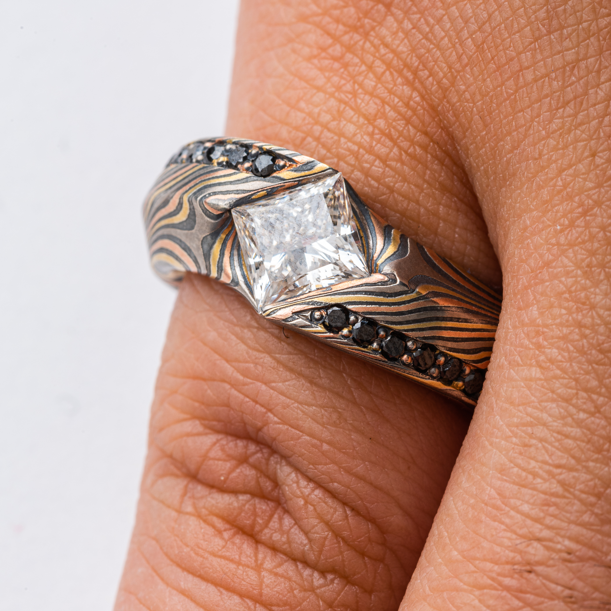 This is a custom made Mokume Gane ring, made by arn krebs, featuring a unique carved angled profile and a modified cathedral style setting holding the large white center diamond. The diamond is square and set diagonally to the band, and is flanked on each side by seven small bead set black diamonds running parallel to the band. It's made in our Twist pattern and an upgraded 18kt version of our Firestorm palette, the metals in it are red gold yellow gold palladium and silver