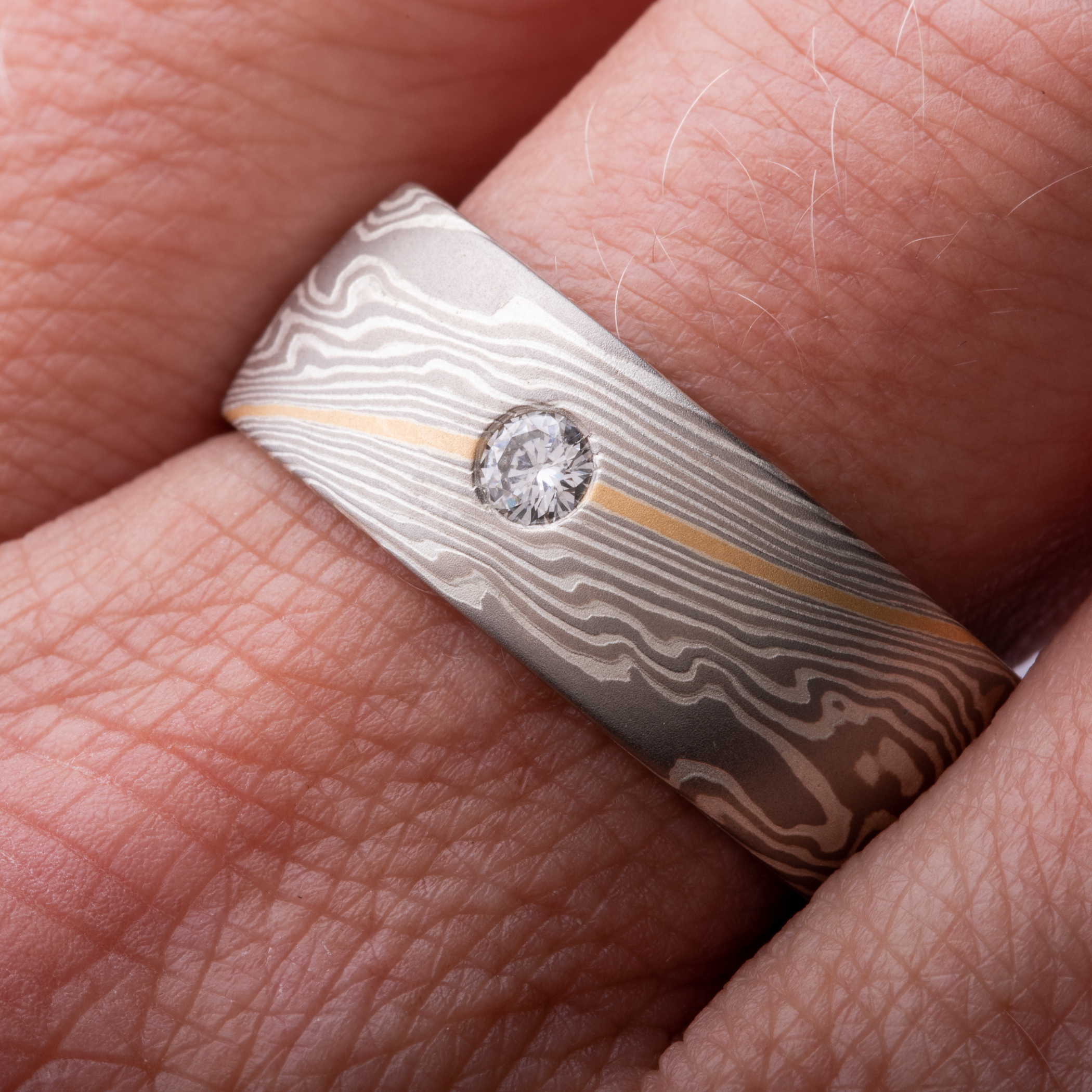 Wide mokume gane men's wedding ring, with a single round diamond flush set into the band, the ring is overall made of layers of gray metals, with one stripe of gold.