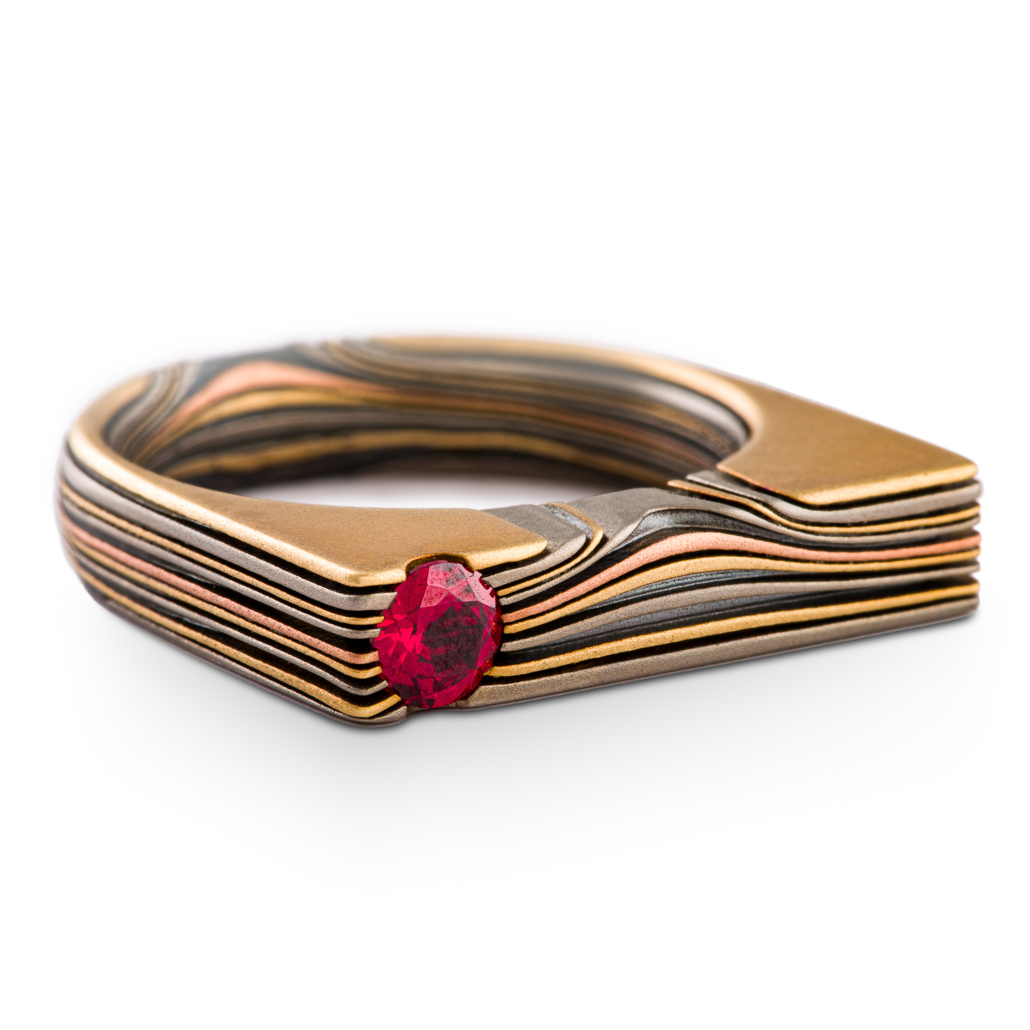 Uniquely shaped flat top mokume gane ring with red stone set flush towards one side. The ring is rounded on the underside for comfort, and made with layers of red and yellow gold, palladium and oxidized silver.