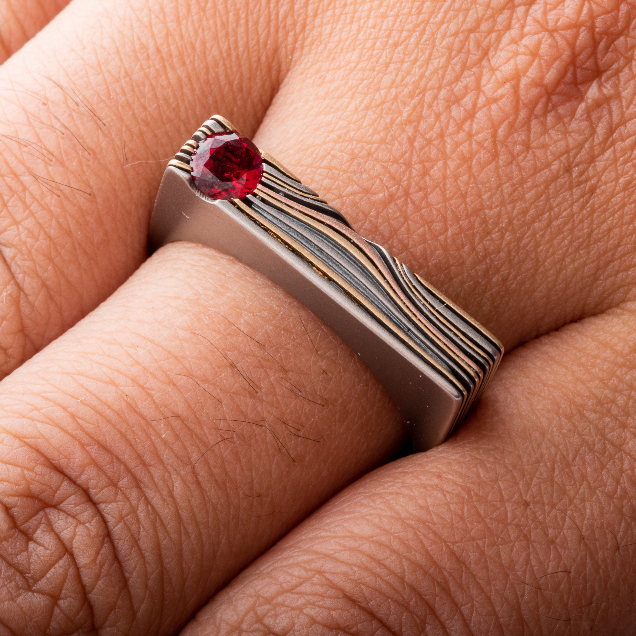 Uniquely shaped flat top mokume gane ring with red stone set flush towards one side. The ring is rounded on the underside for comfort, and made with layers of red and yellow gold, palladium and oxidized silver.