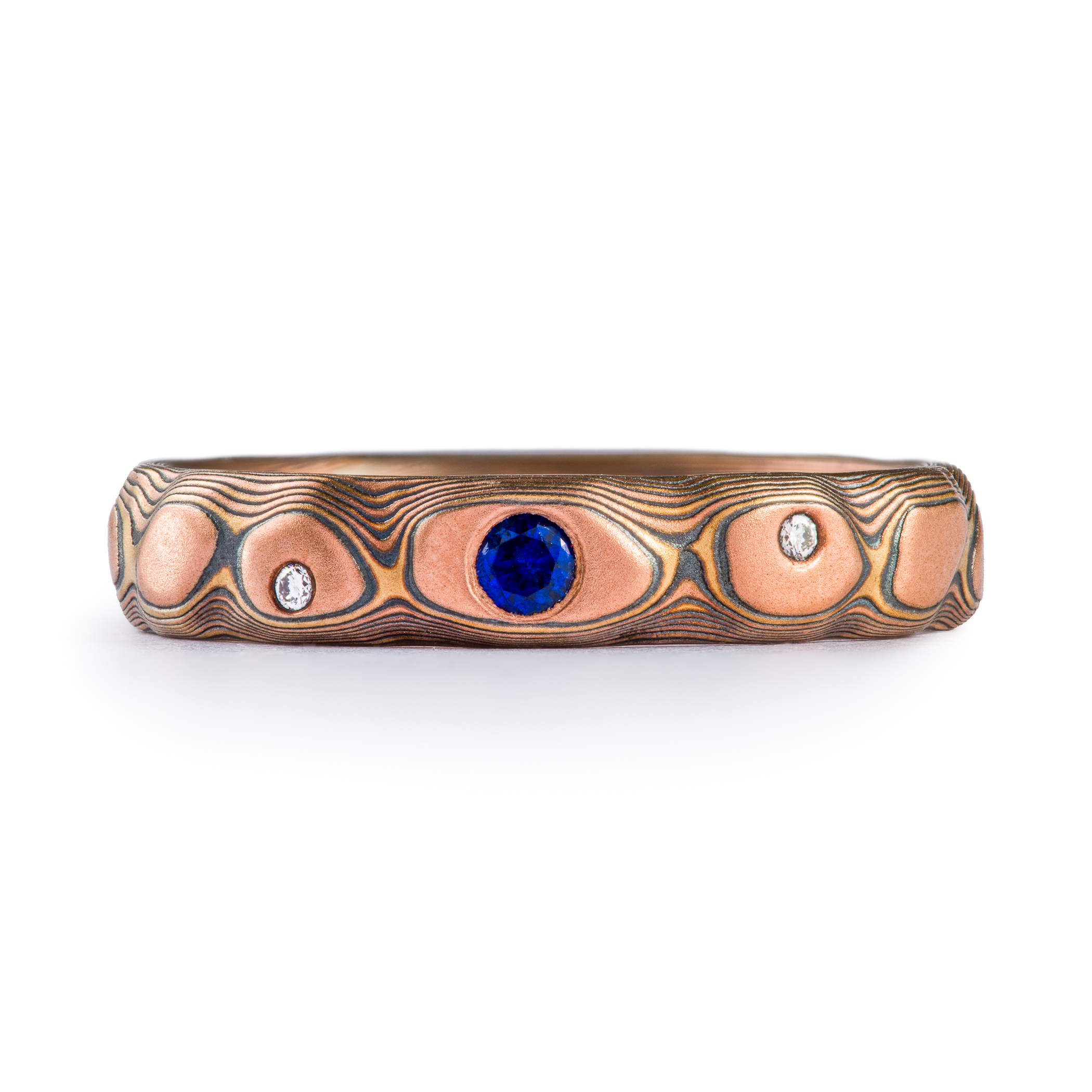 Textured red gold mokume gane ring, with three flush set stones, a slightly larger blue sapphire, flanked by small white stones.