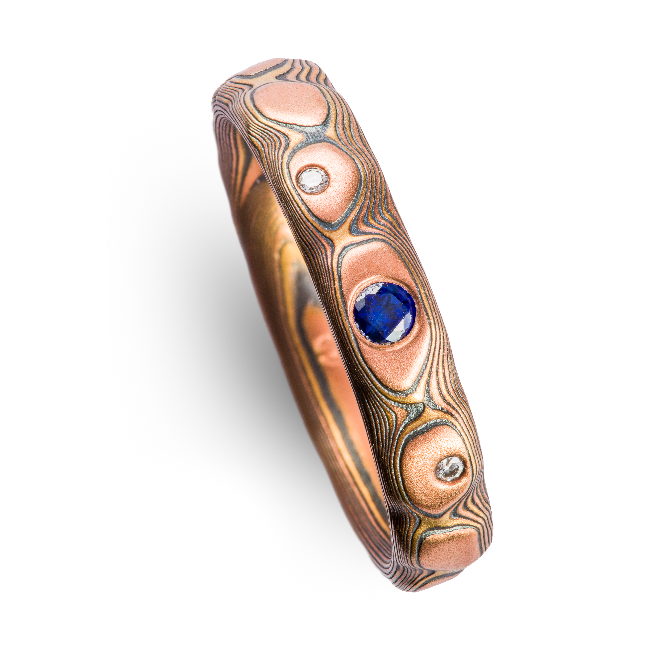 Textured red gold mokume gane ring, with three flush set stones, a slightly larger blue sapphire, flanked by small white stones.