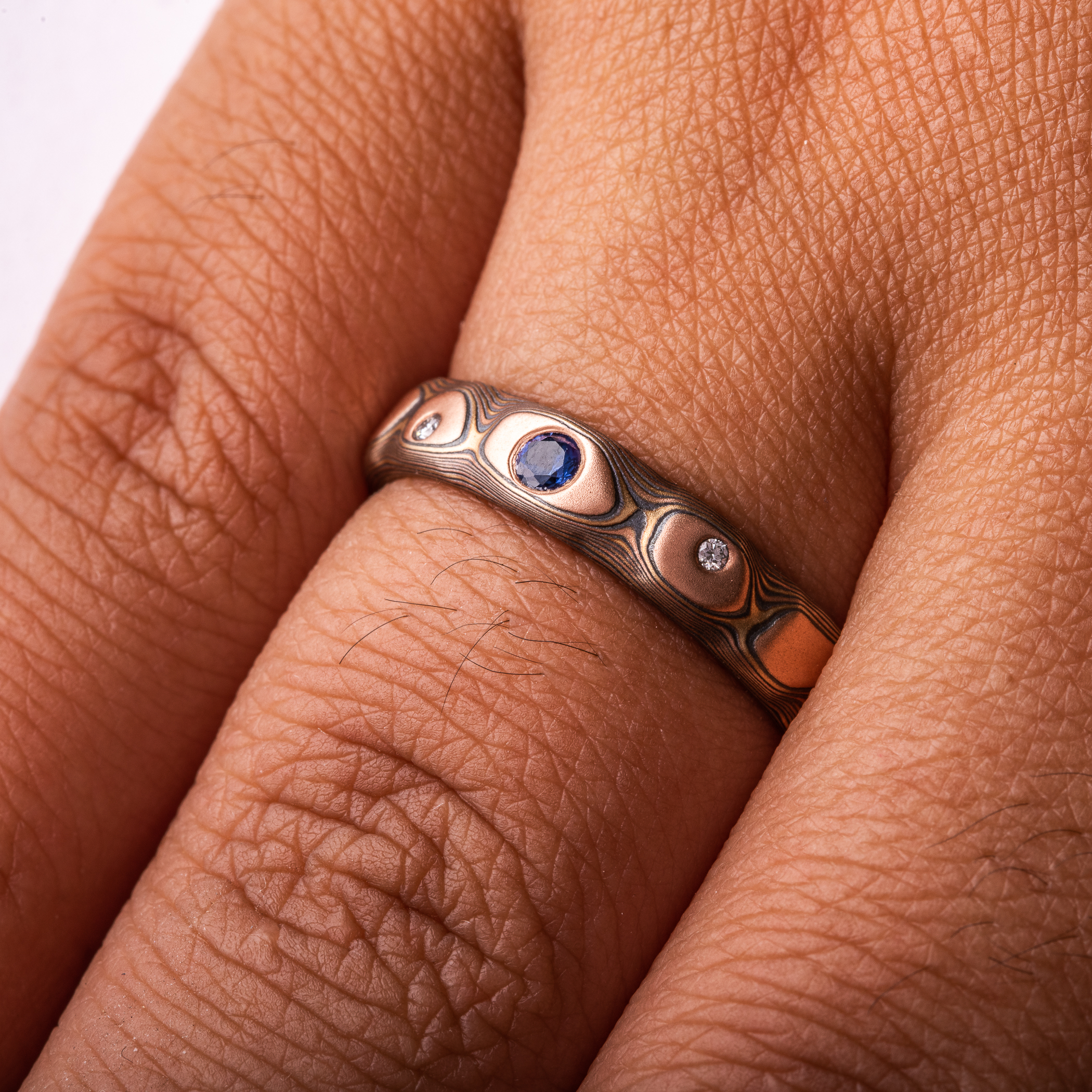 Textured red gold mokume gane ring, with three flush set stones, a slightly larger blue sapphire, flanked by small white stones.
