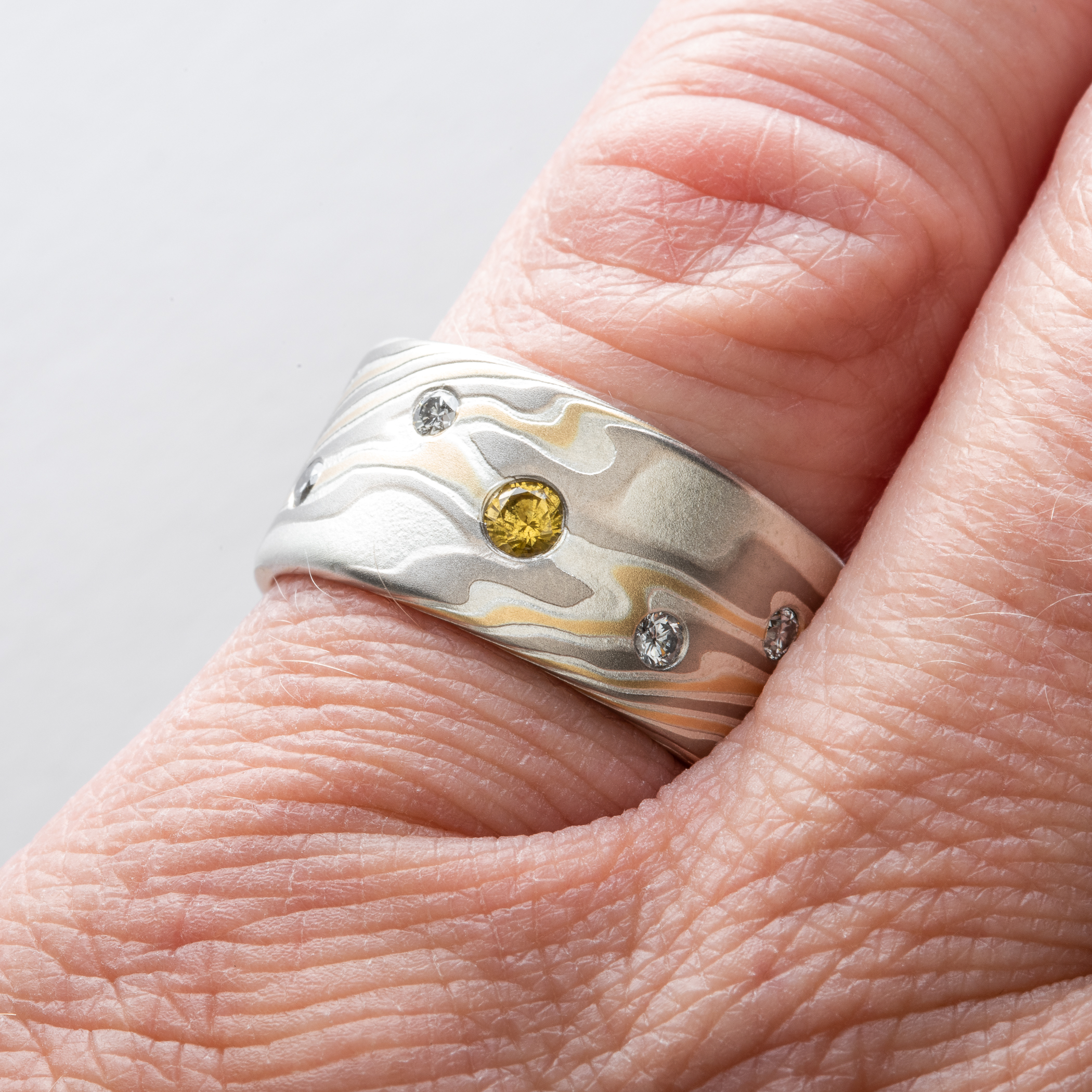 Mokume gane patterned ring made with yellow gold, palladium and silver, with small round yellow and white stones set into the wide, flat profile band.