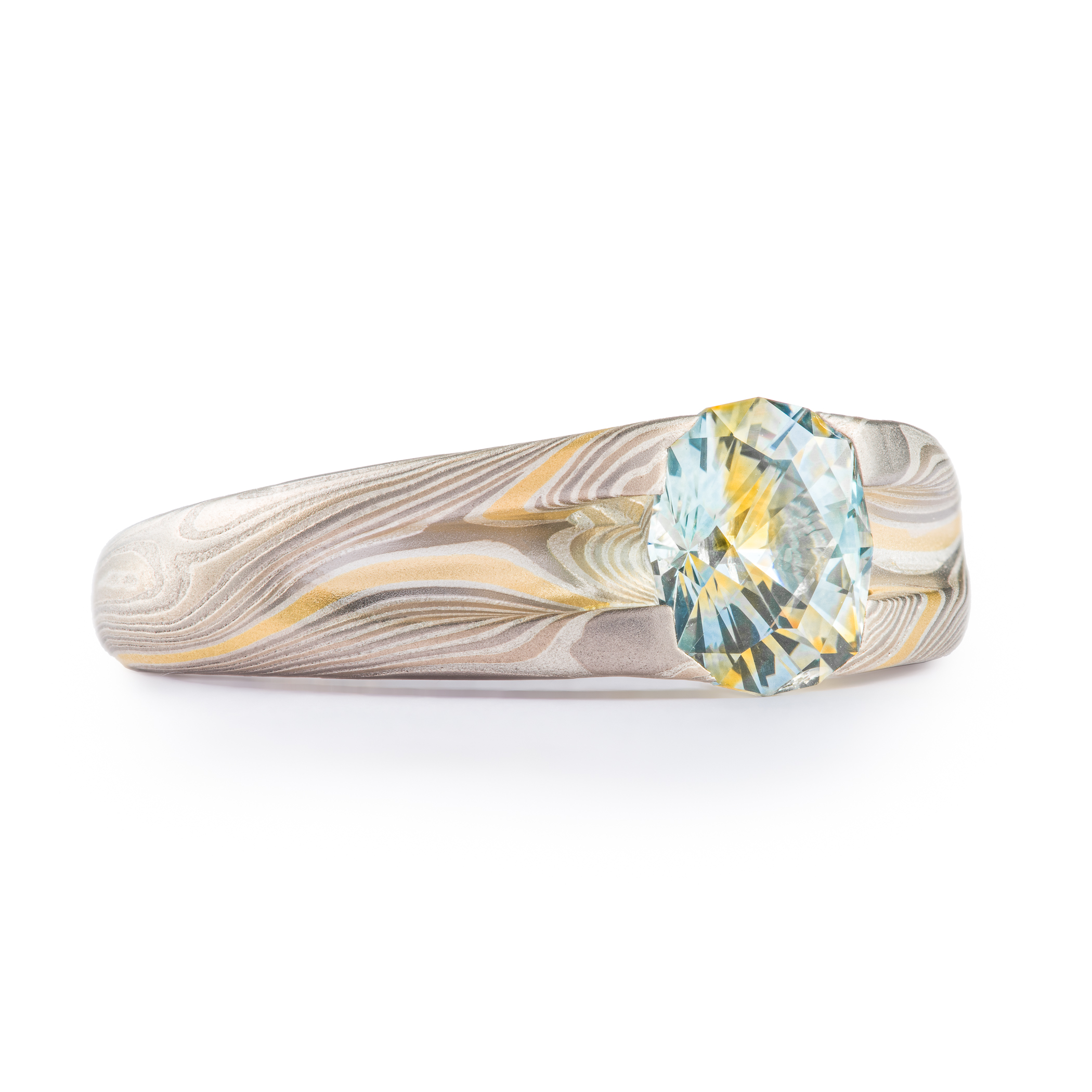 Mokume gane patterned ring with large multicolored sapphire held in prongs carved from the body of the ring. The stone is a light blue color, with a gold stripe running through the center, echoing the gold stripe running throughout the rest of the band.
