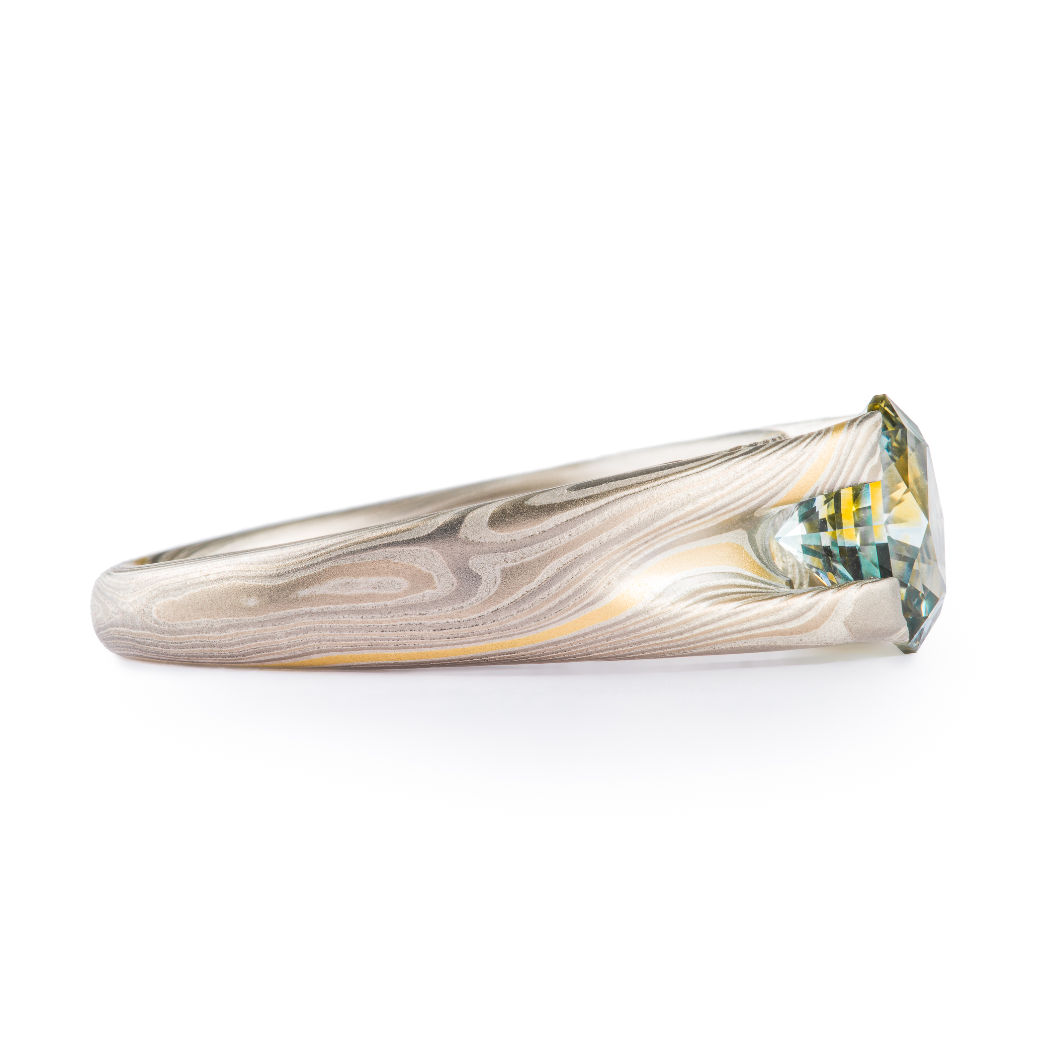 Mokume gane patterned ring with large multicolored sapphire held in prongs carved from the body of the ring. The stone is a light blue color, with a gold stripe running through the center, echoing the gold stripe running throughout the rest of the band.