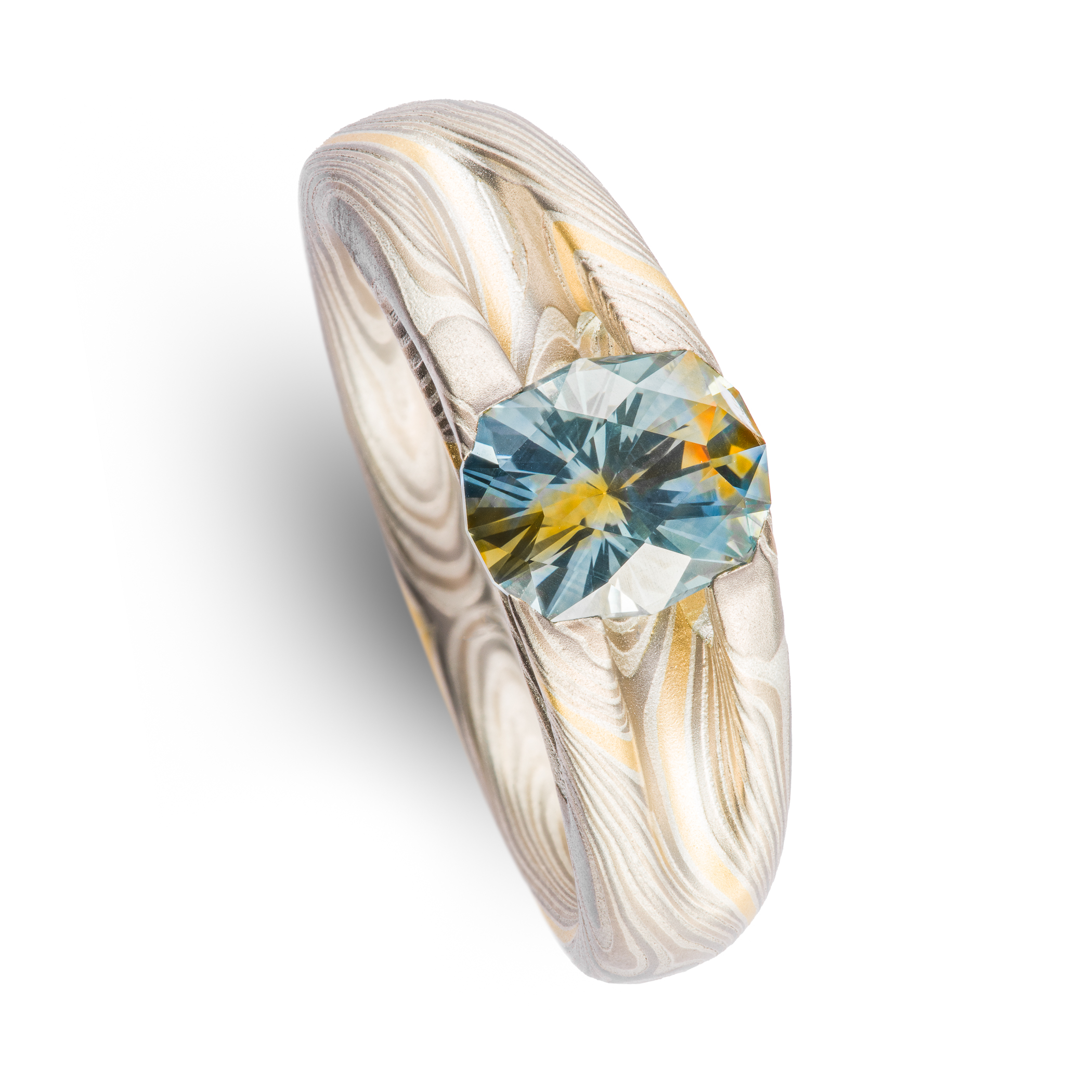 Mokume gane patterned ring with large multicolored sapphire held in prongs carved from the body of the ring. The stone is a light blue color, with a gold stripe running through the center, echoing the gold stripe running throughout the rest of the band.