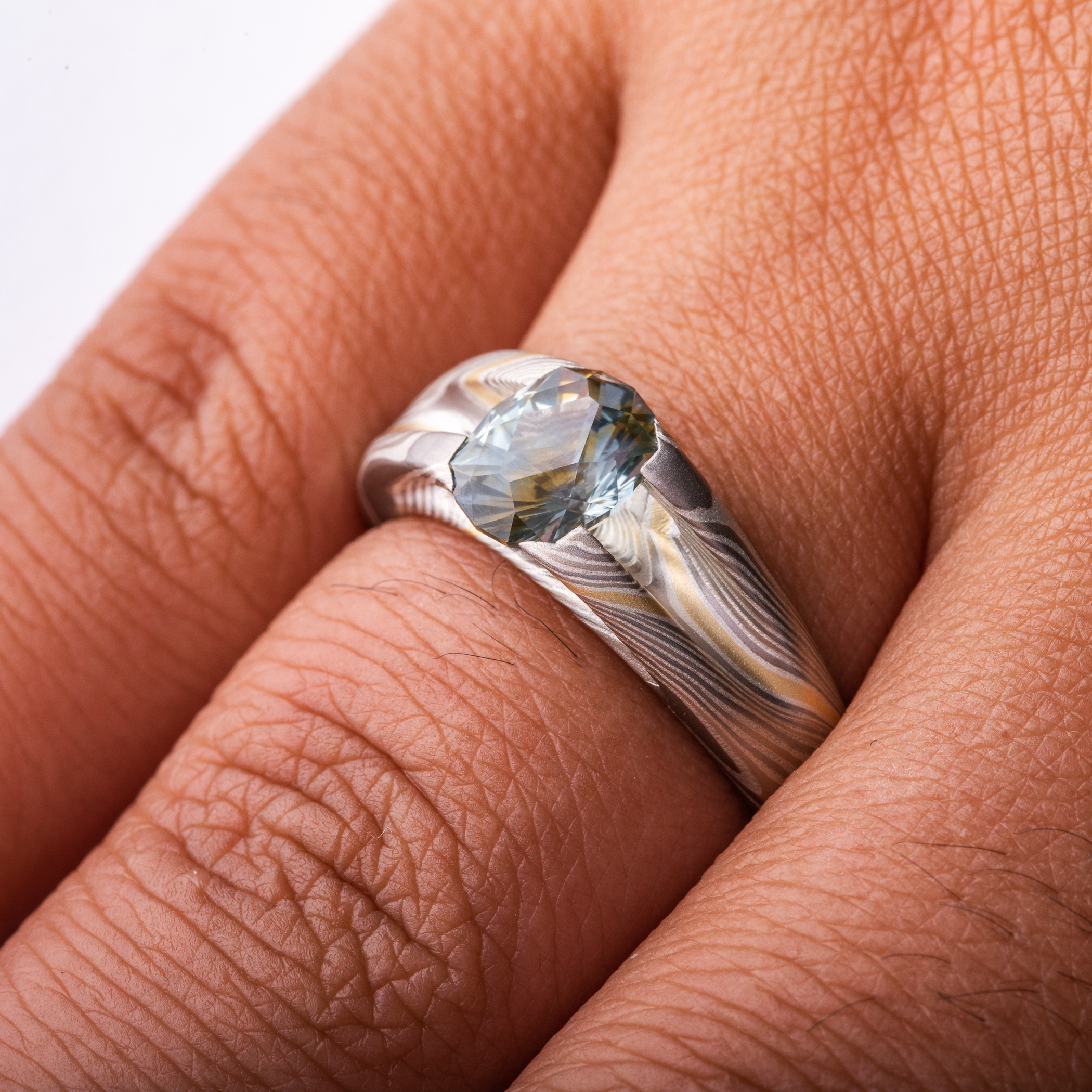 Mokume gane patterned ring with large multicolored sapphire held in prongs carved from the body of the ring. The stone is a light blue color, with a gold stripe running through the center, echoing the gold stripe running throughout the rest of the band.