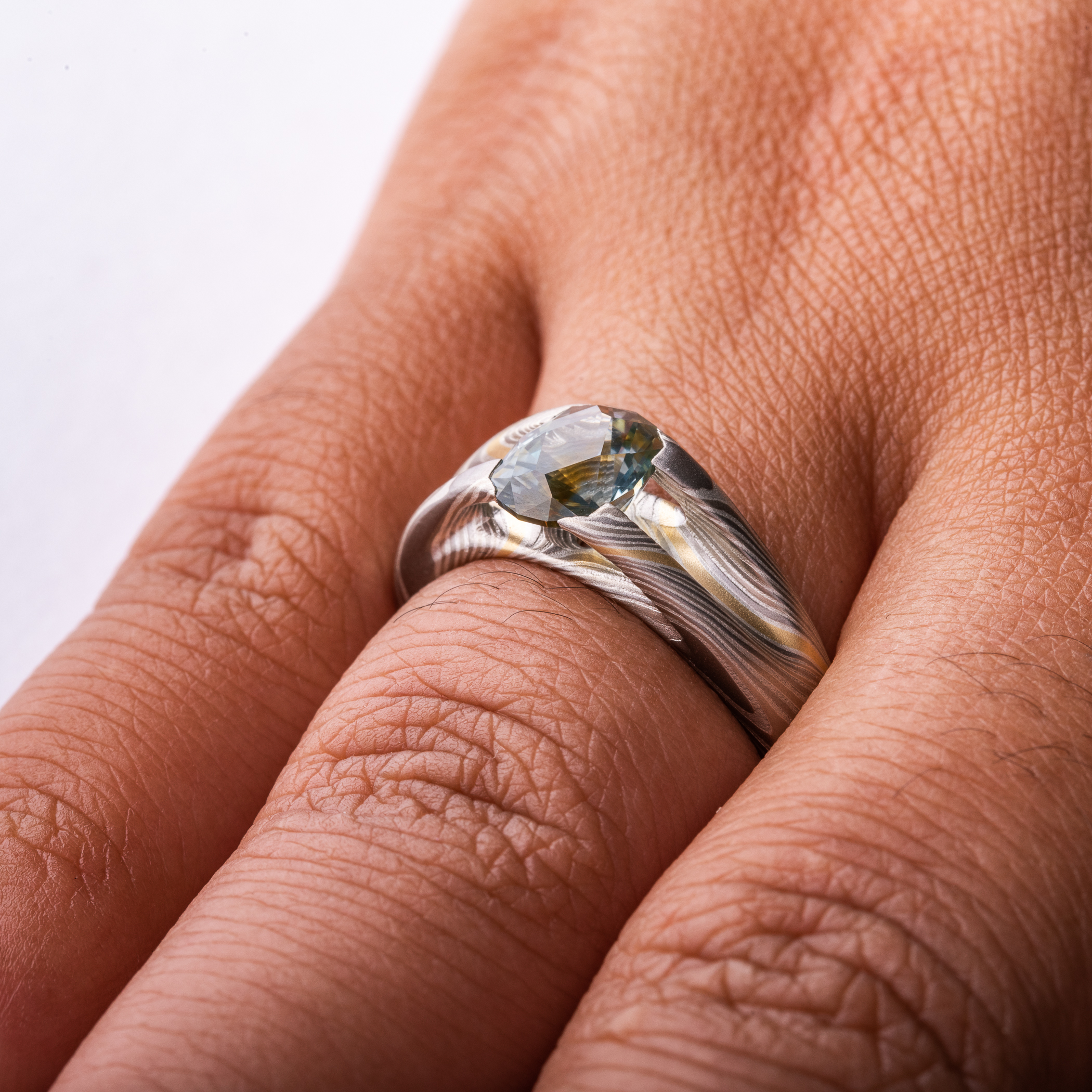 Mokume gane patterned ring with large multicolored sapphire held in prongs carved from the body of the ring. The stone is a light blue color, with a gold stripe running through the center, echoing the gold stripe running throughout the rest of the band.