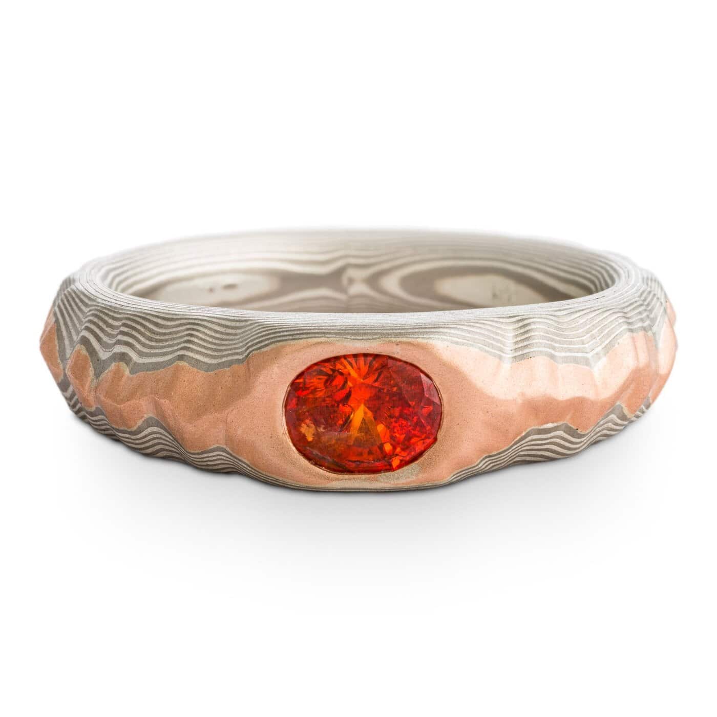 Mokume gane ring with oval deep orange sapphire, the ring is patterned to look like a mountain range, the main ring is gray and silver, topped with red gold.