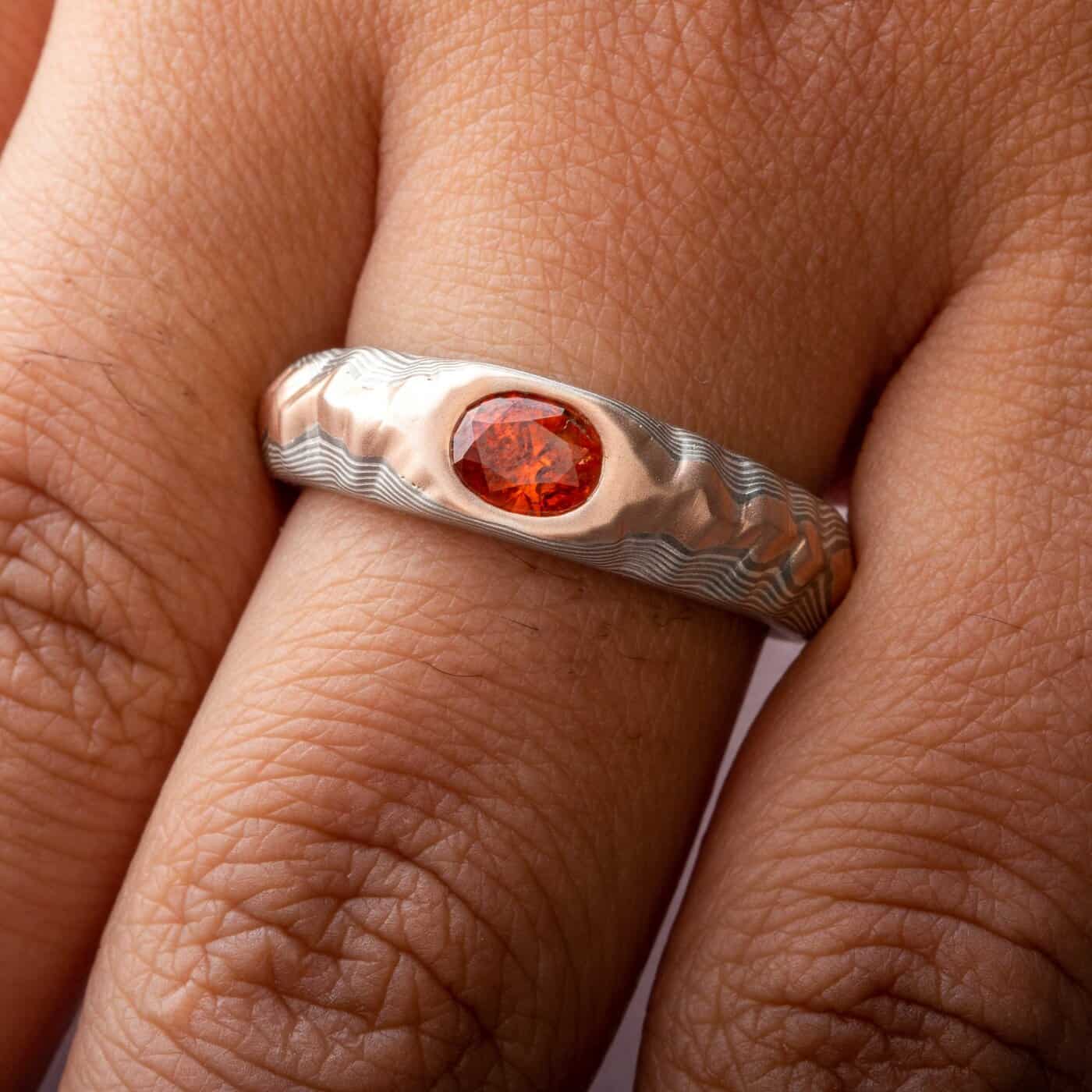 Mokume gane ring with oval deep orange sapphire, the ring is patterned to look like a mountain range, the main ring is gray and silver, topped with red gold.