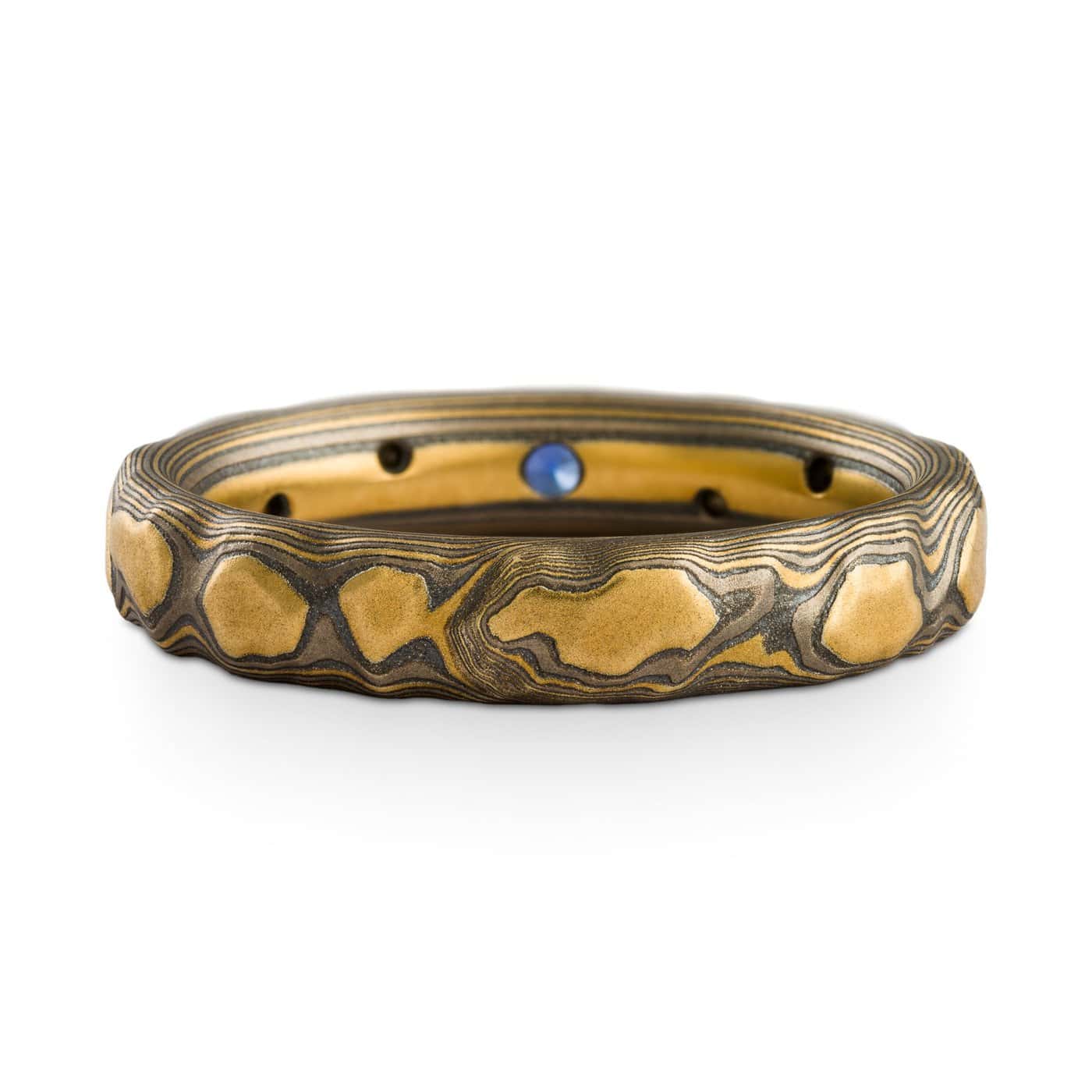 Narrow mokume gane ring made with yellow gold, palladium and oxidized silver, in a three dimensional pattern made to resemble rocks. The ring has small flush set stones, one larger blue one at the center, and smaller white stones placed organically throughout.