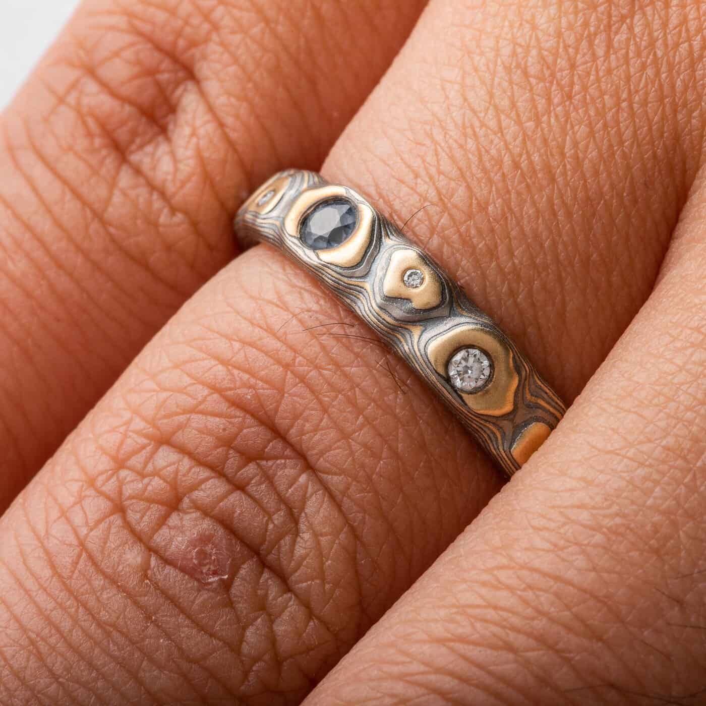 Narrow mokume gane ring made with yellow gold, palladium and oxidized silver, in a three dimensional pattern made to resemble rocks. The ring has small flush set stones, one larger blue one at the center, and smaller white stones placed organically throughout.
