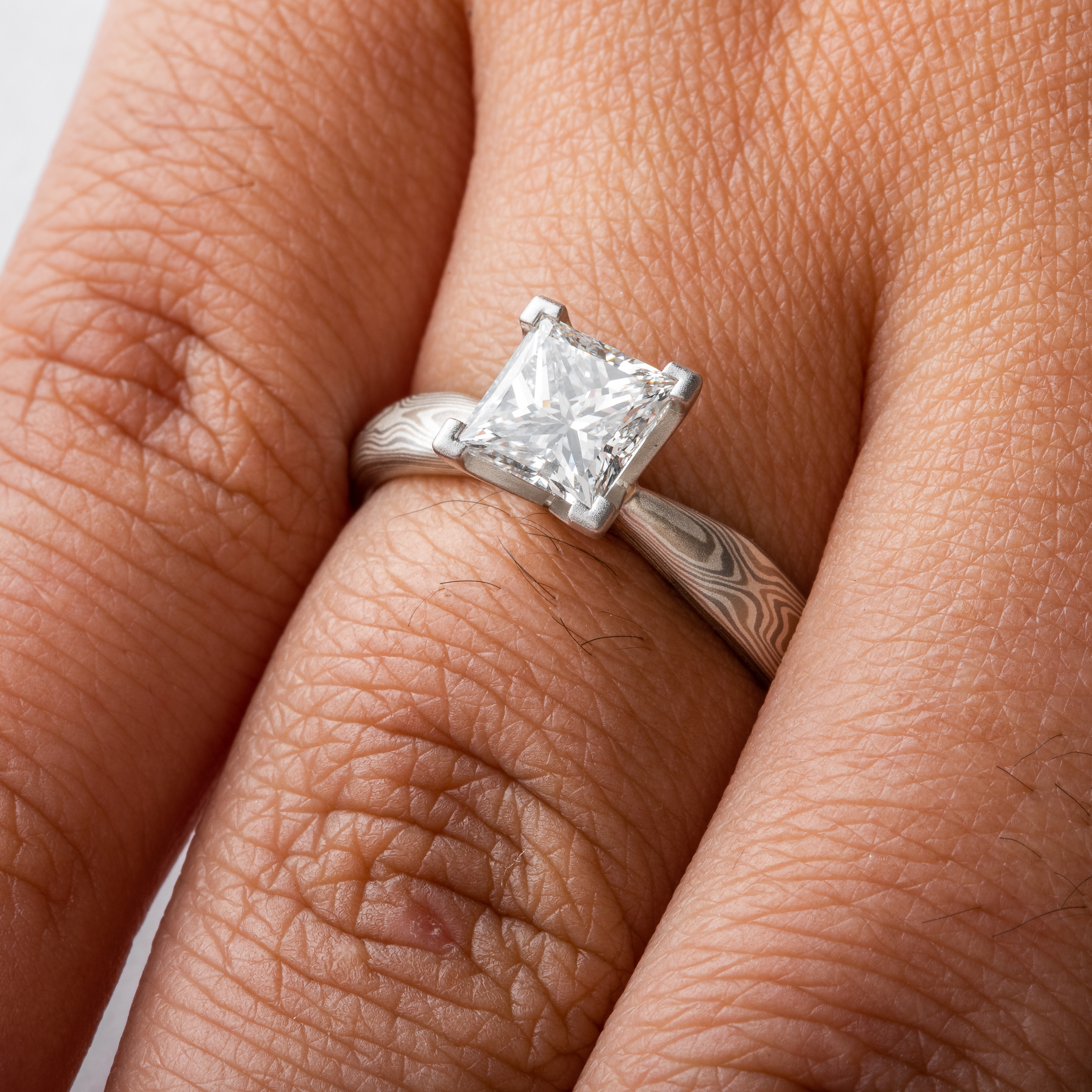 solitaire style mokume gane engagement ring with square diamond set in prongs, ring has an overall silvery palette and a woodgrain style