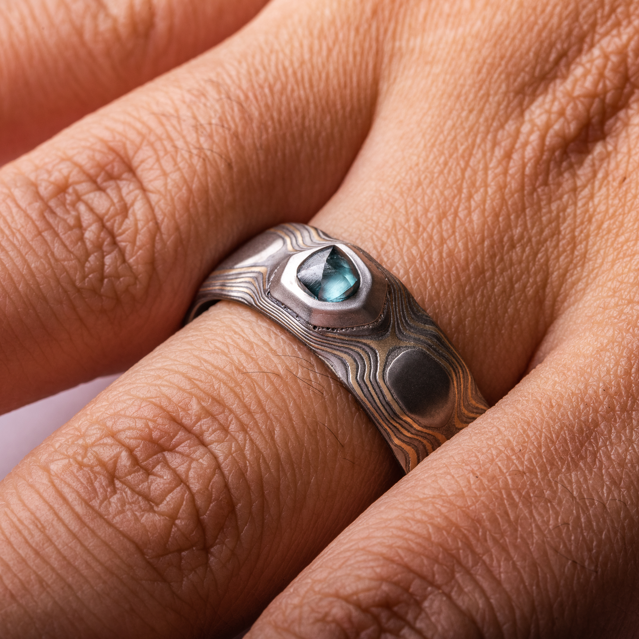 Wide men's style mokume gane ring carved to look like rocks, with a blue diamond crystal set into it, looking like it's emerging from the earth.