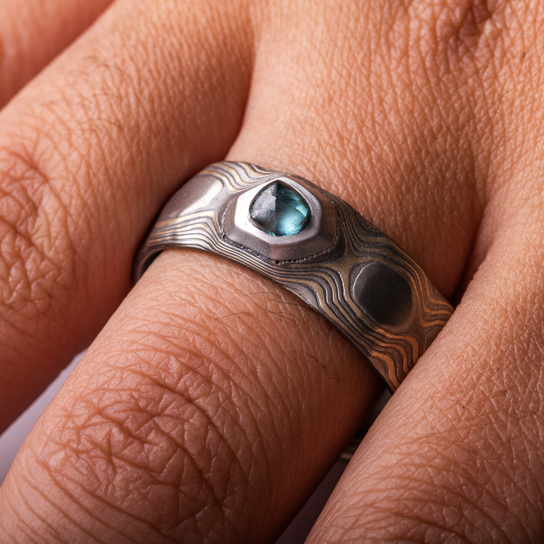 Wide men's style mokume gane ring carved to look like rocks, with a blue diamond crystal set into it, looking like it's emerging from the earth.