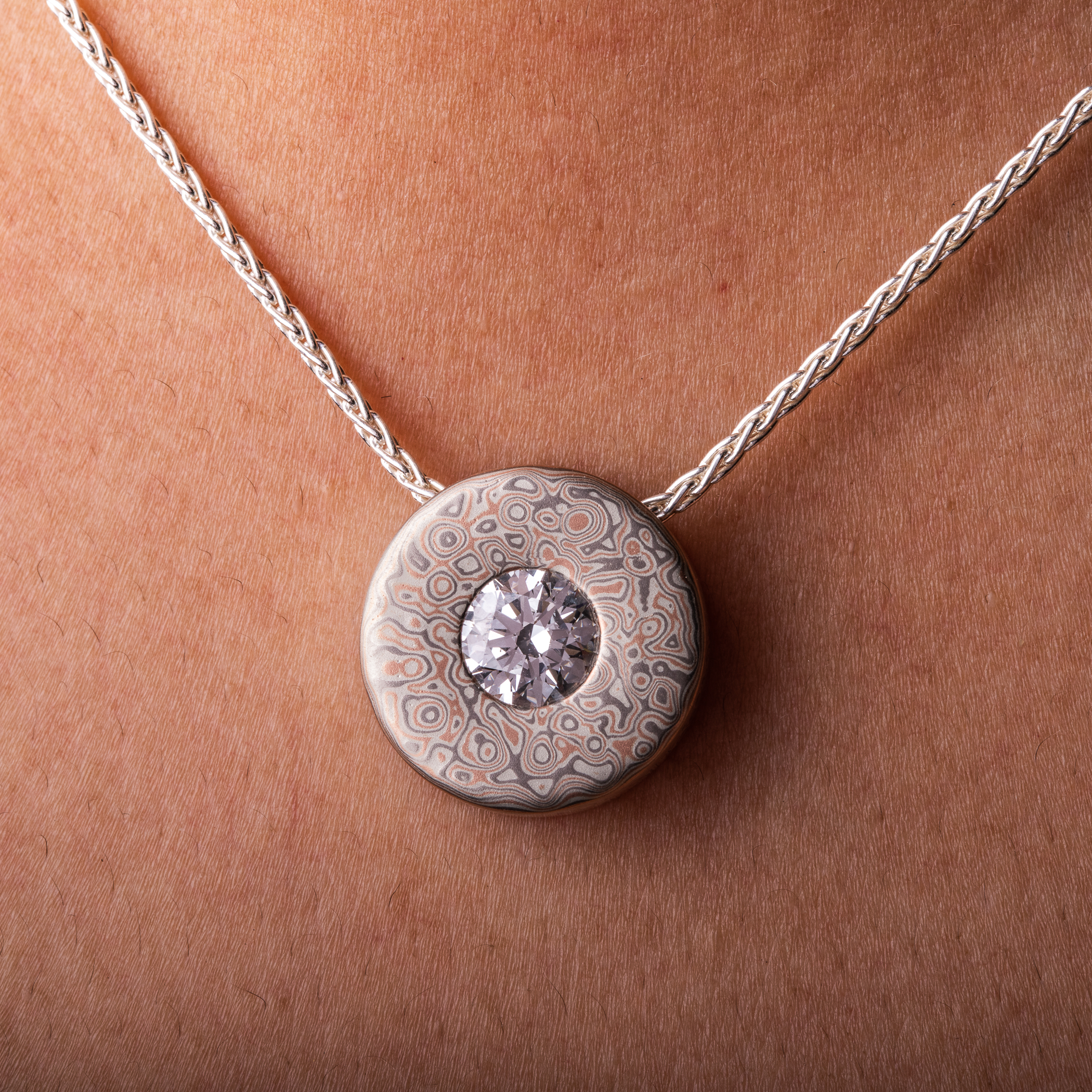 Small round pendant with a moissanite set in the center on a chain, patterned in a style that looks like raindrops on water.
