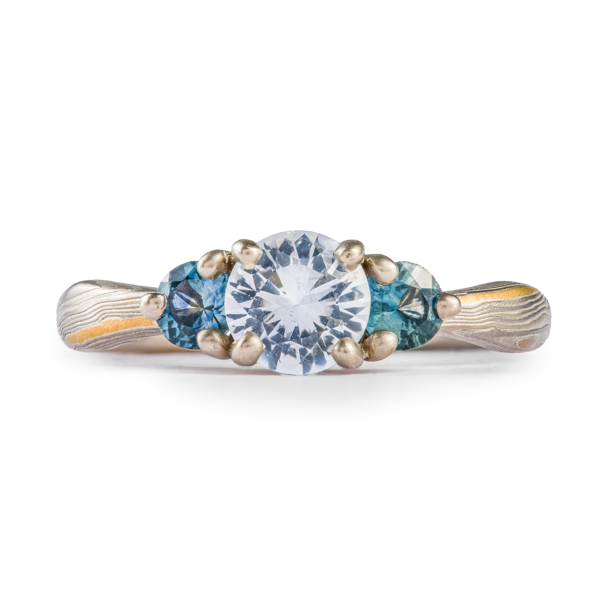mokume gane engagement ring with a white center stone and two smaller blue stones on either side, set in prongs.