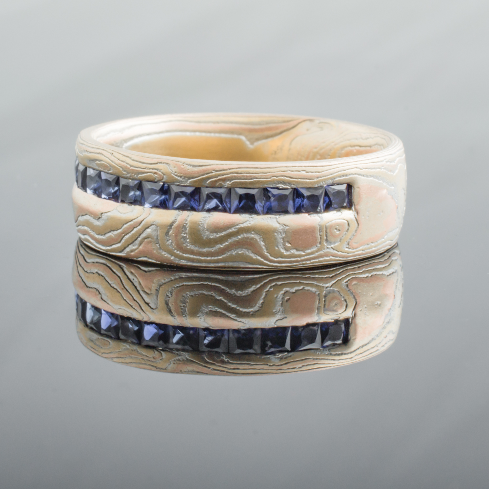 Mokume gane woodgrain patterned ring made with red and yellow gold, with a line of blue square stones set in a line around the ring towards one edge.
