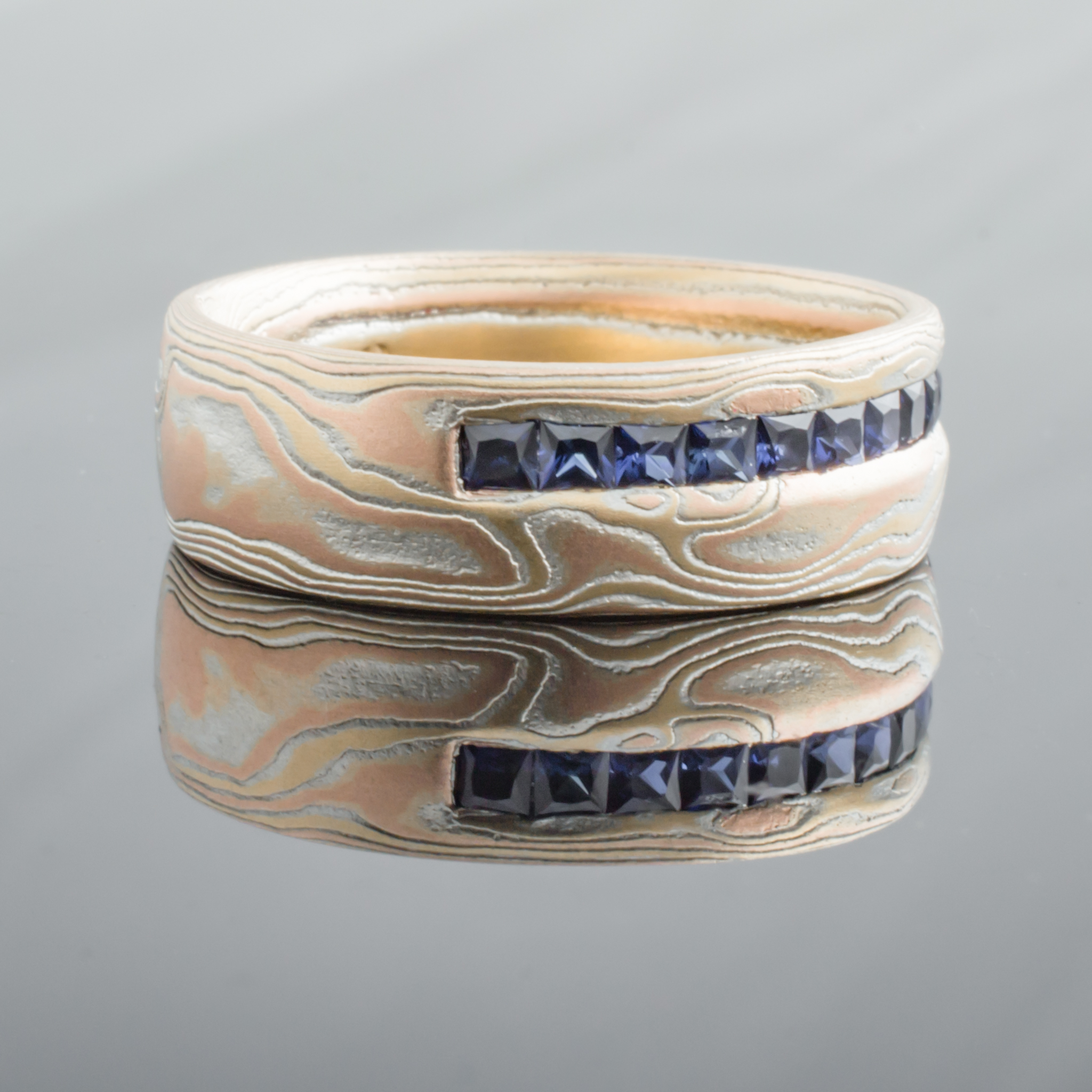 Mokume gane woodgrain patterned ring made with red and yellow gold, with a line of blue square stones set in a line around the ring towards one edge.