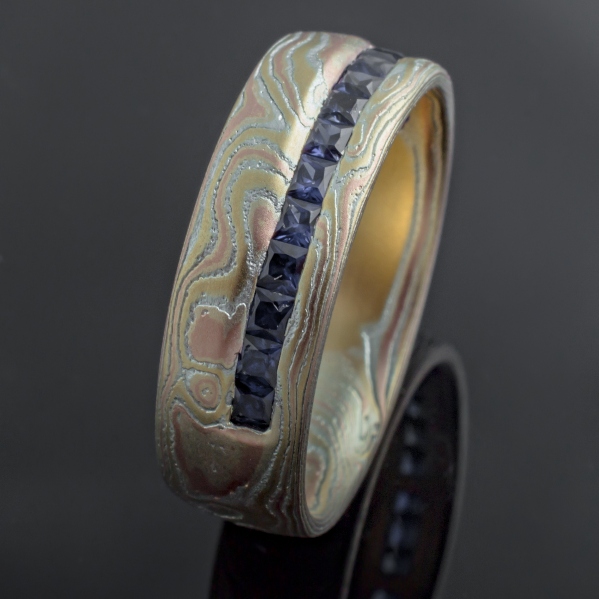 Mokume gane woodgrain patterned ring made with red and yellow gold, with a line of blue square stones set in a line around the ring towards one edge.