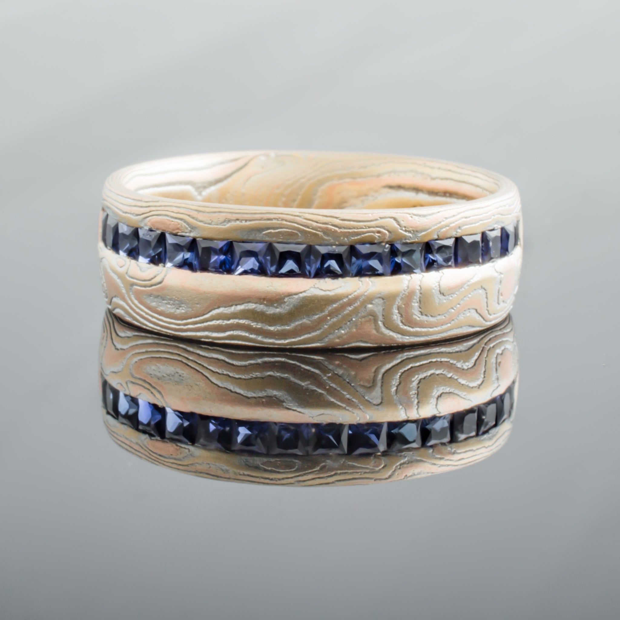 Mokume gane woodgrain patterned ring made with red and yellow gold, with a line of blue square stones set in a line around the ring towards one edge.