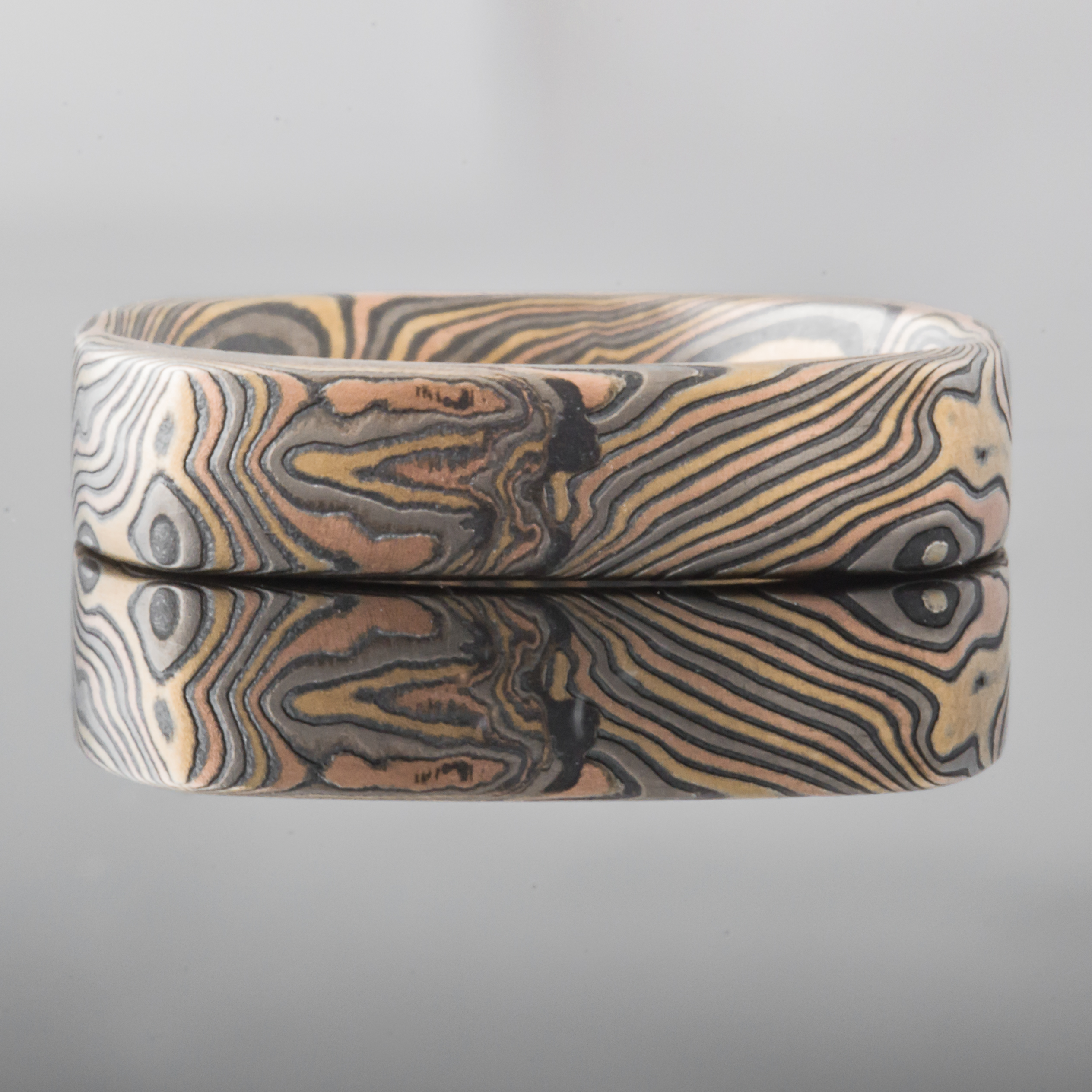 Mokume gane patterned ring in a unique soft square shape, with a small red stone flush set towards one side.