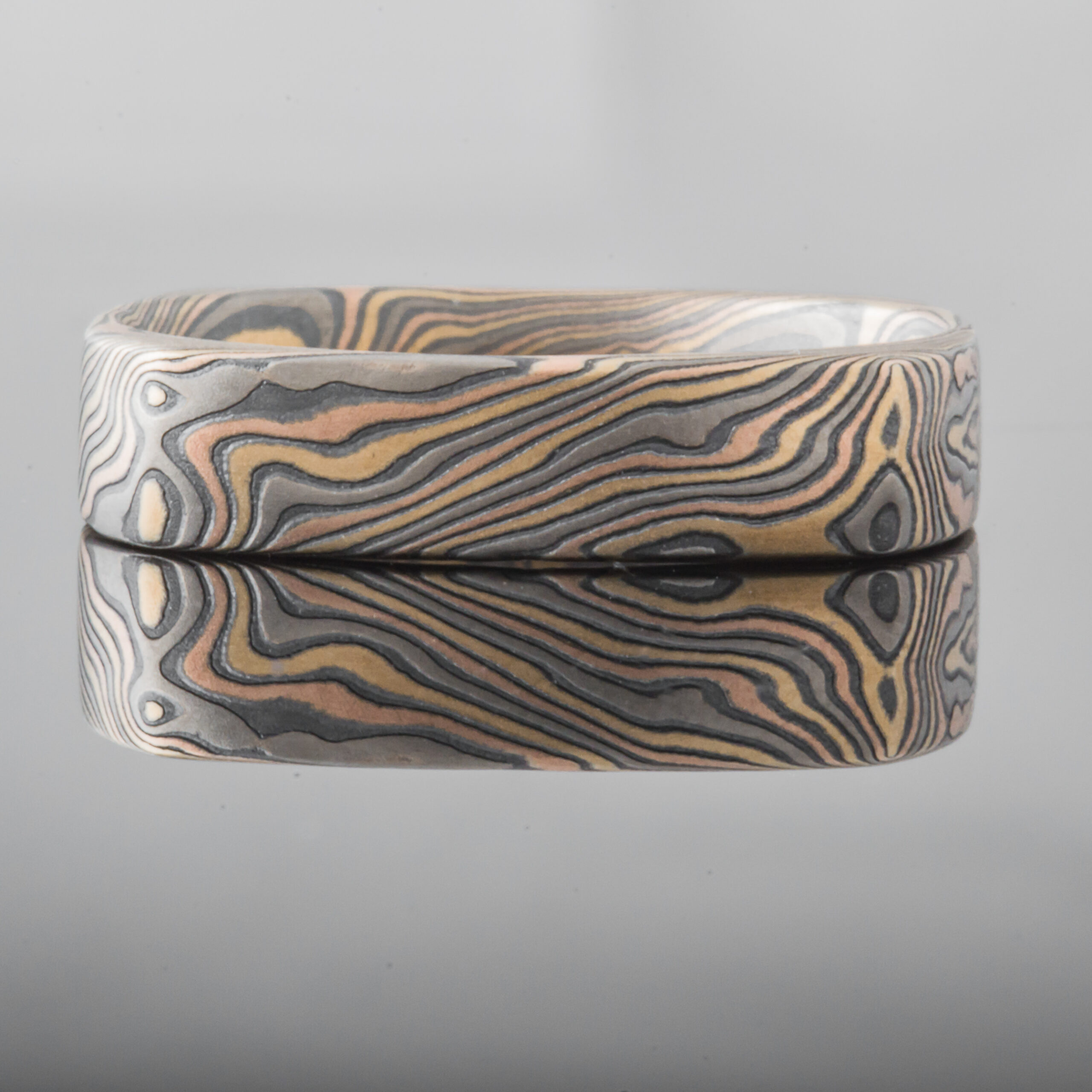 Mokume gane patterned ring in a unique soft square shape, with a small red stone flush set towards one side.
