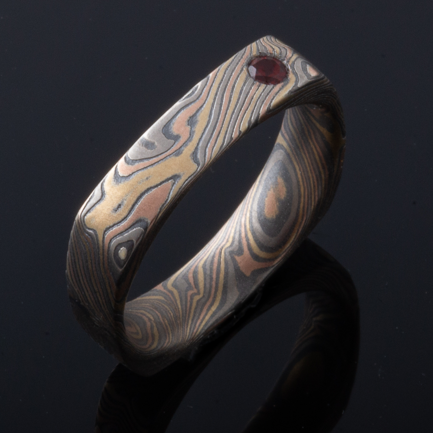 Mokume gane patterned ring in a unique soft square shape, with a small red stone flush set towards one side.