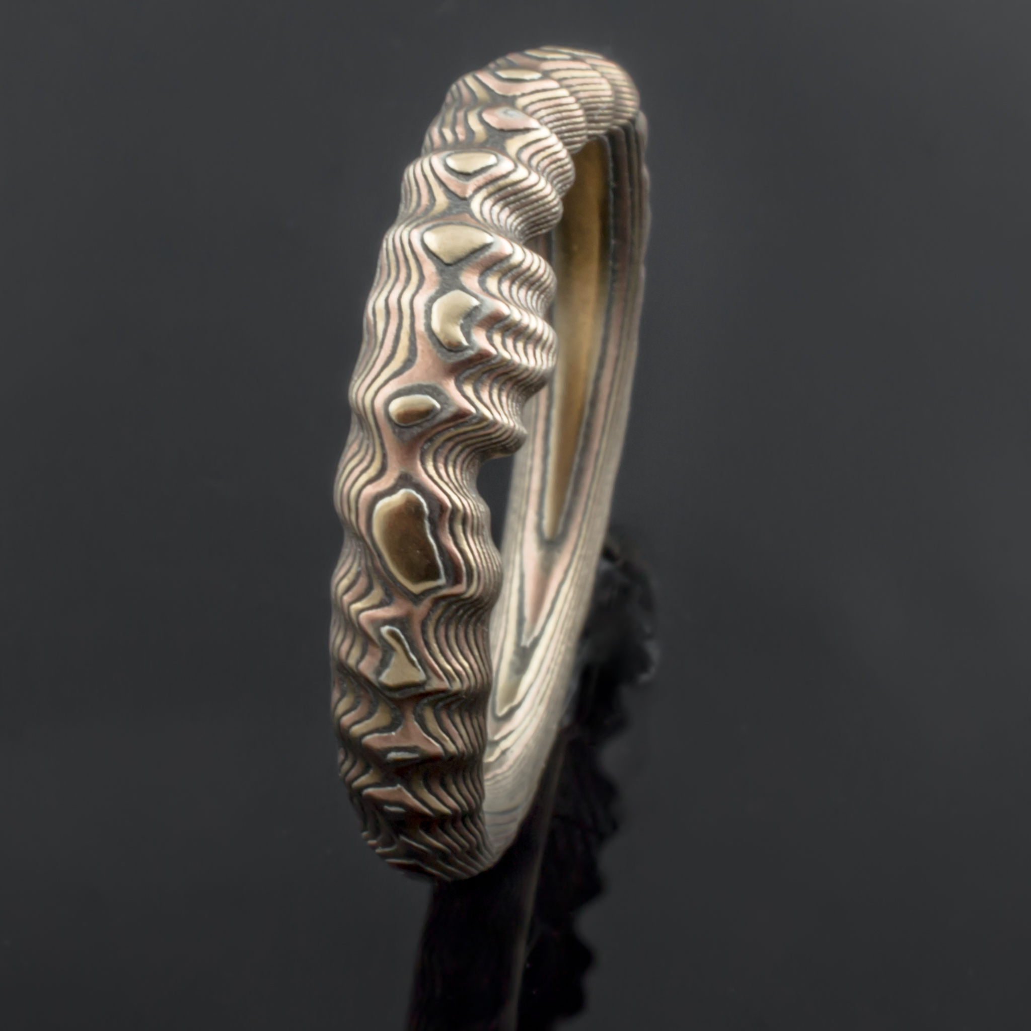 Uniquely contoured mokume gane ring with a shaped edge made to fit with an engagement ring, and textured to look like topography.