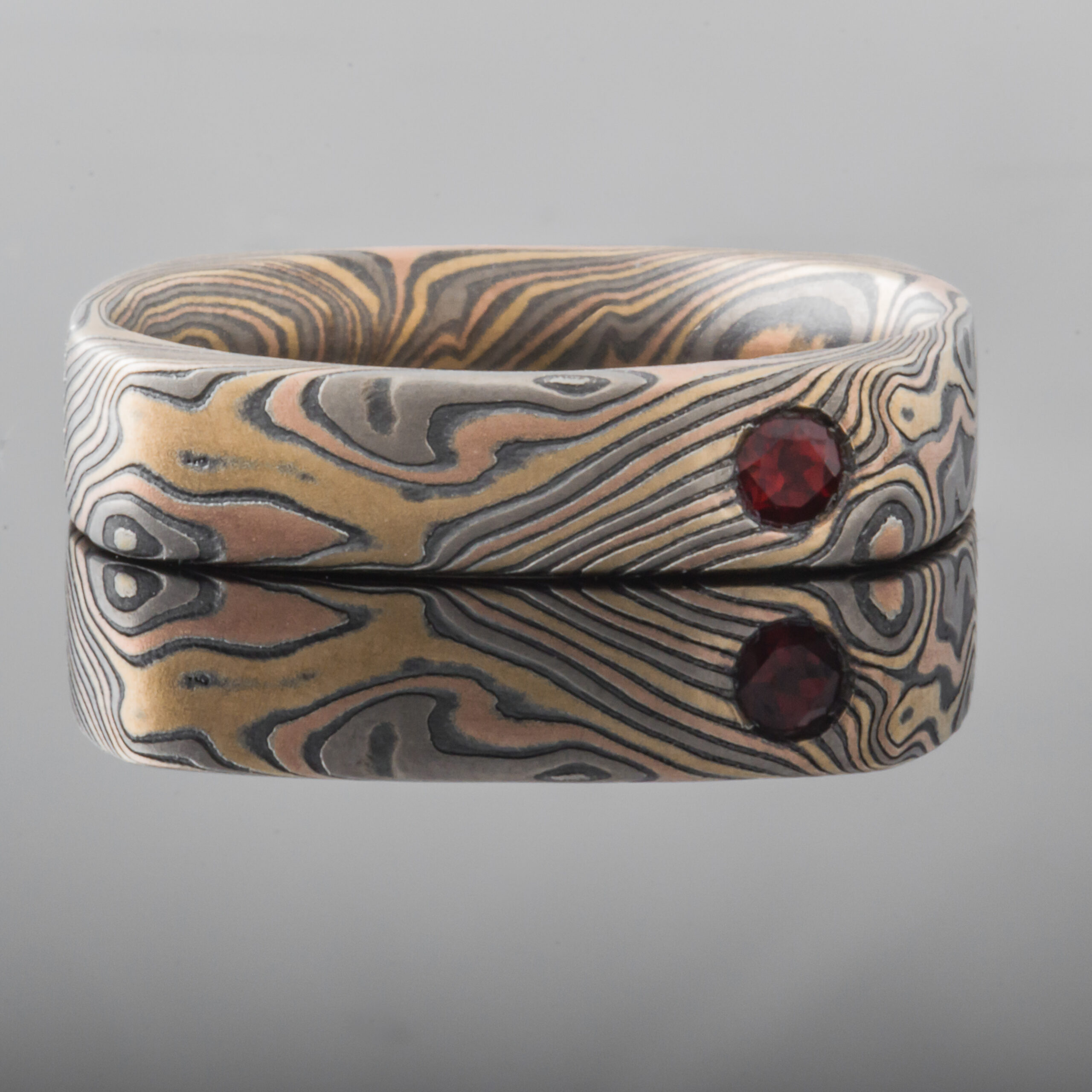 Mokume gane patterned ring in a unique soft square shape, with a small red stone flush set towards one side.