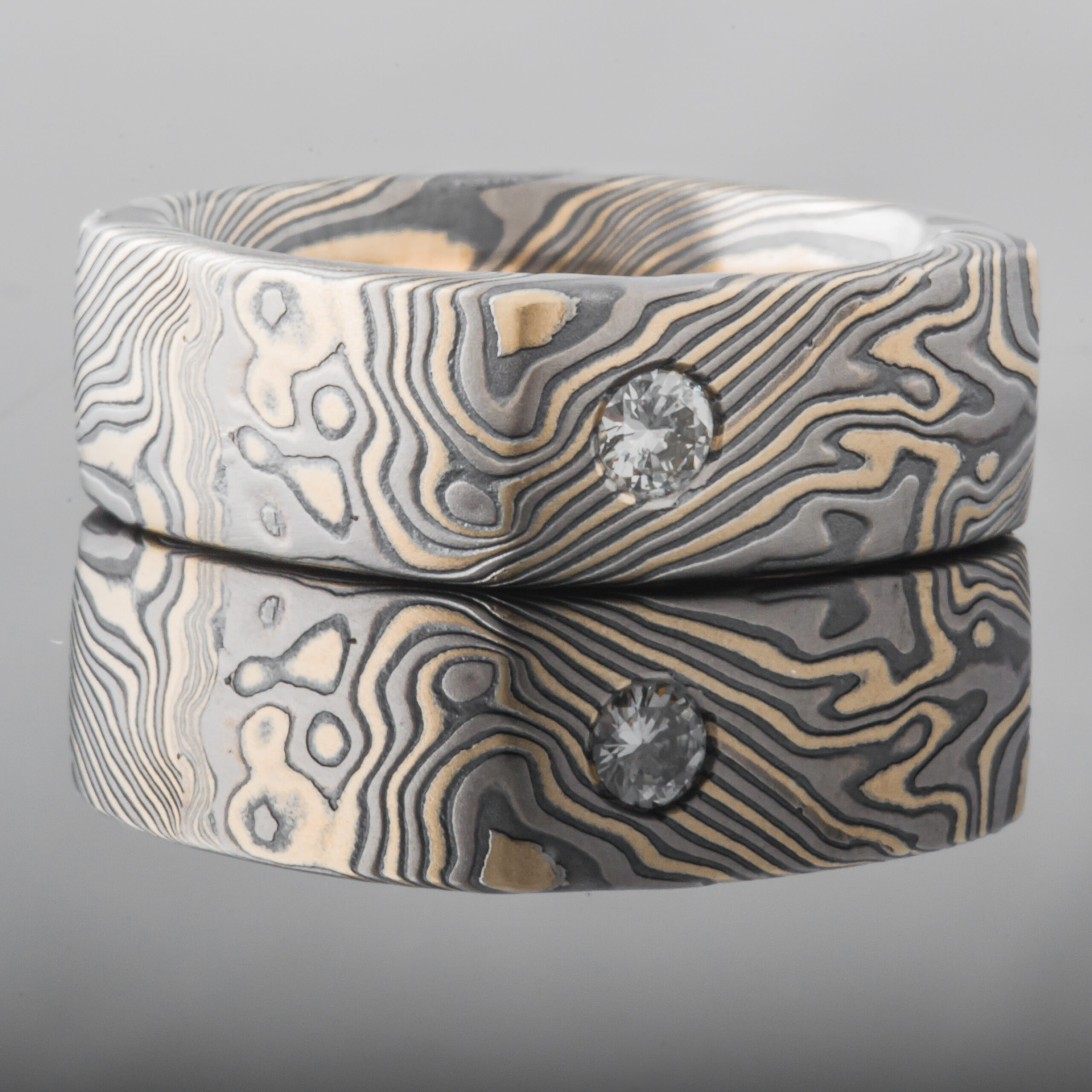 Wide rustic men's style square mokume gane ring made with yellow gold, palladium and oxidized silver, with a round white stone flush set into the band.