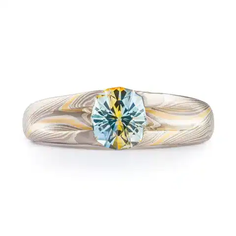 bold statement style mokume gane ring with a custom carved prong setting holding a large multicolor sapphire, the sapphire is primarily blue with a streak of yellow running through the center