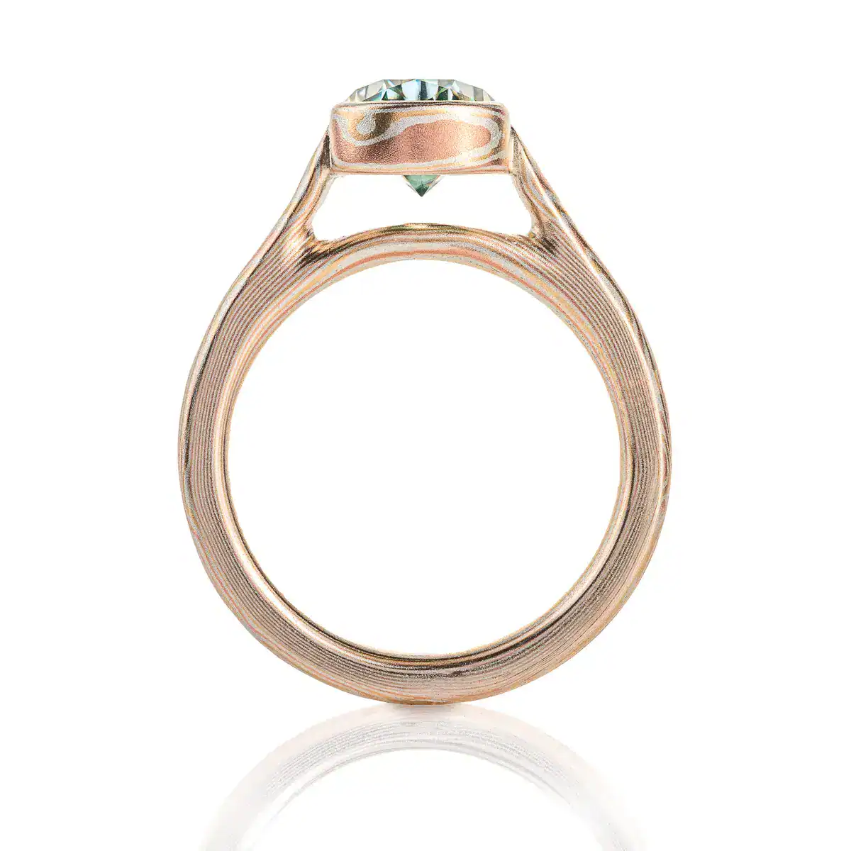 mokume gane ring with green moissanite in a pear shape, the stone is held in a mokume bezel, bezel and ring are patterned in woodgrain, with red gold yellow gold and silver