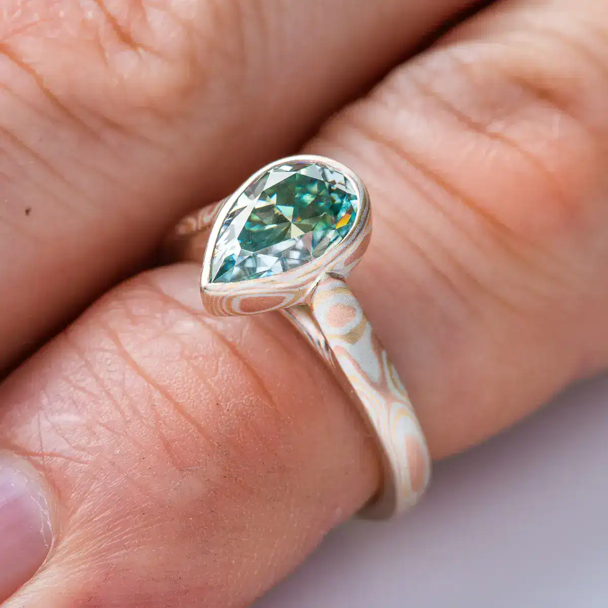 mokume gane ring with green moissanite in a pear shape, the stone is held in a mokume bezel, bezel and ring are patterned in woodgrain, with red gold yellow gold and silver