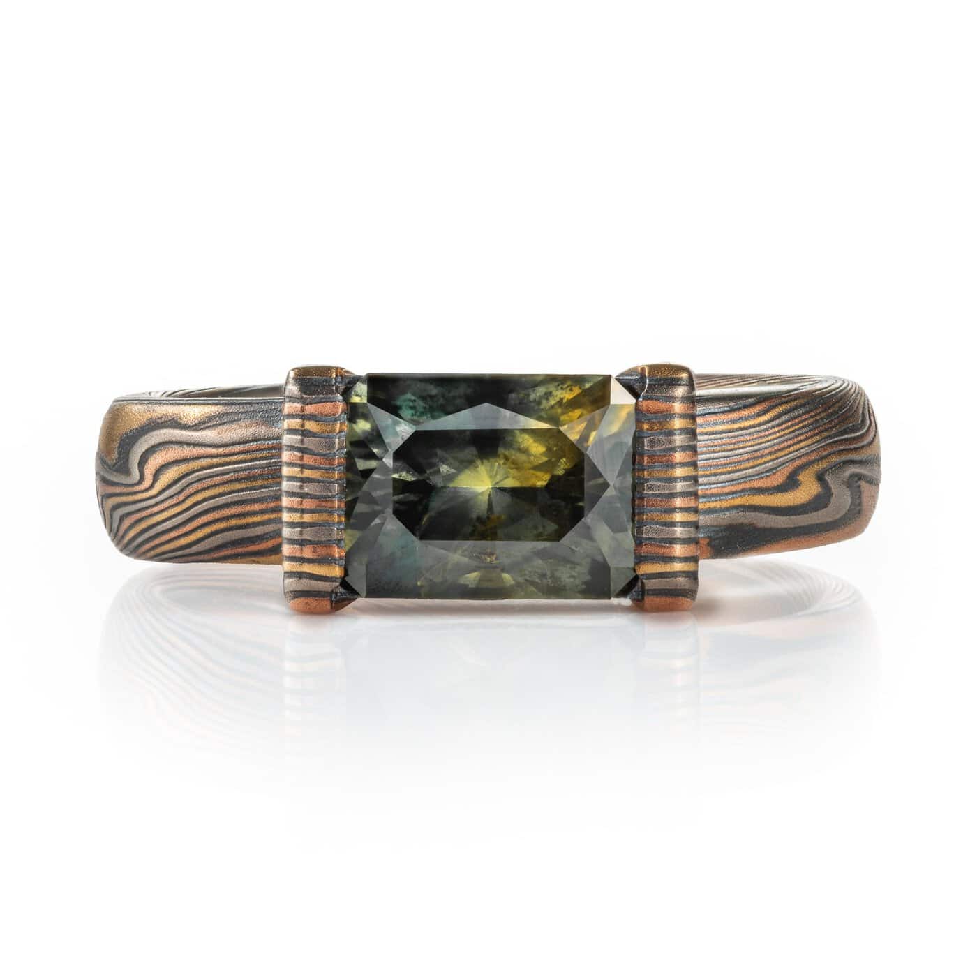 mokume gane ring with larger mossy green rectangular sapphire set horizontally in a partial bezel, with an overall darker palette