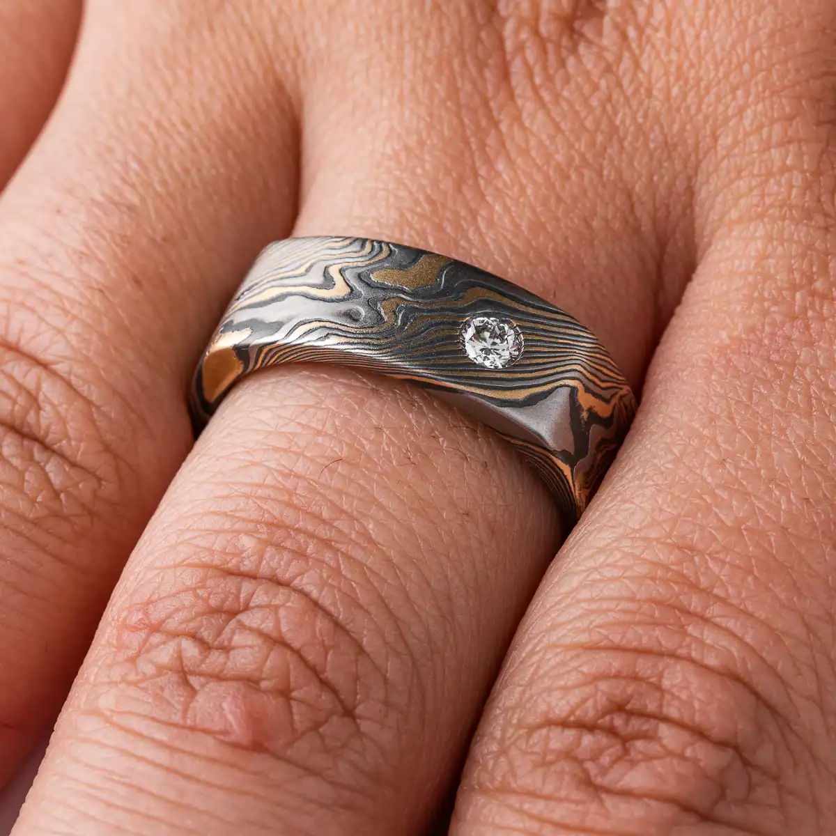 uniquely shaped mokume gane band with a soft square shape on the outside (inside is still circular), with a flat profile for a modern look, and two small stones set flush into the surface, one white and one red.