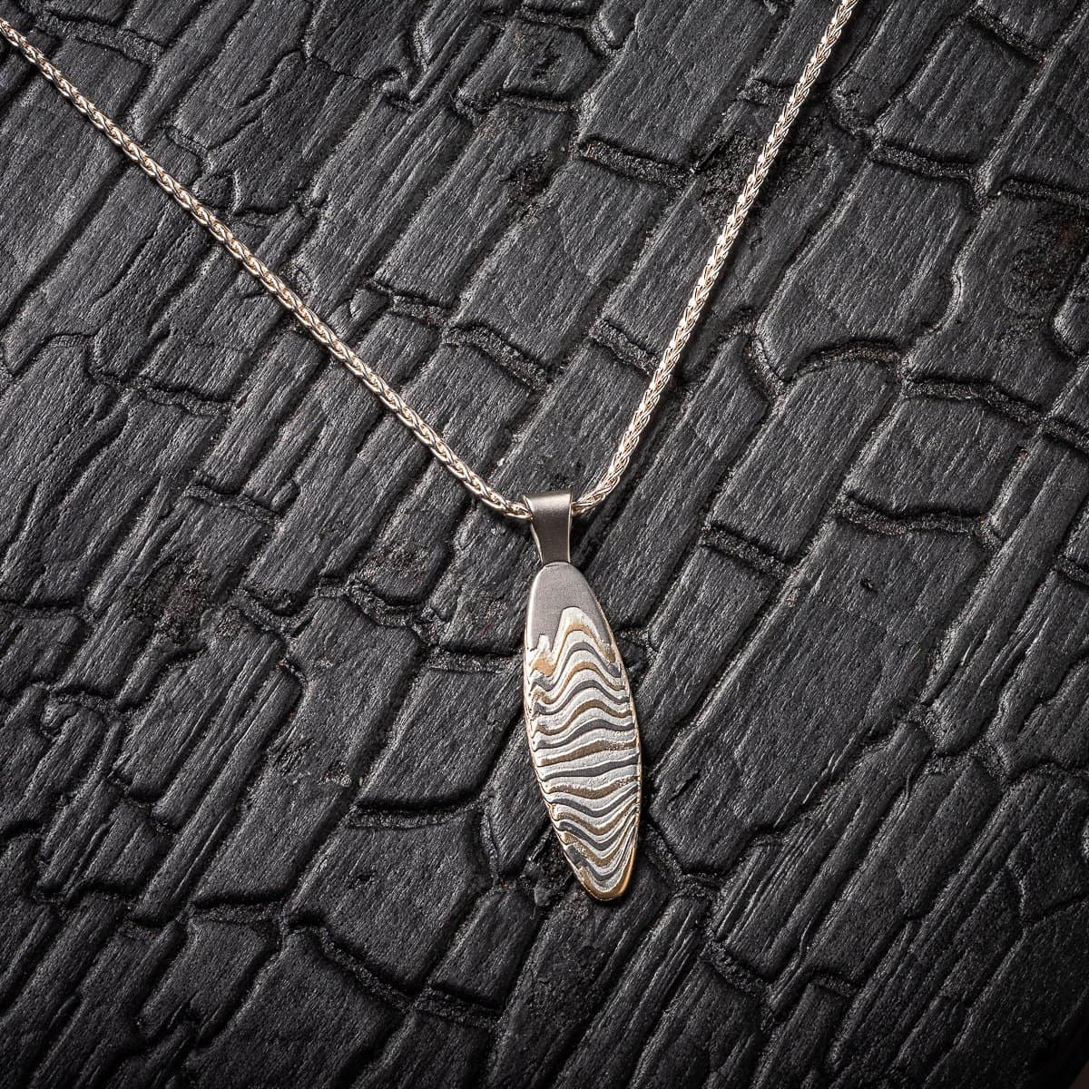 Slim oval shaped mokume gane patterned pendant, made with layers of yellow gold, palladium and silver, on a silver chain. .