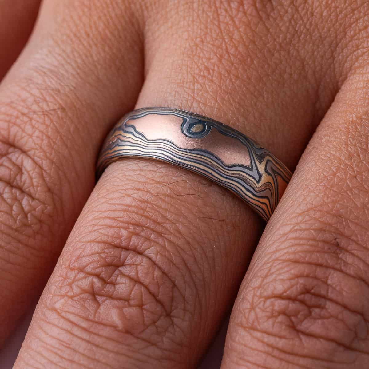 Wide men's style mokume gane band with a domed profile, made in a woodgrain style pattern to resemble wood, with layers of red gold and yellow gold alternating with oxidized silver (black).