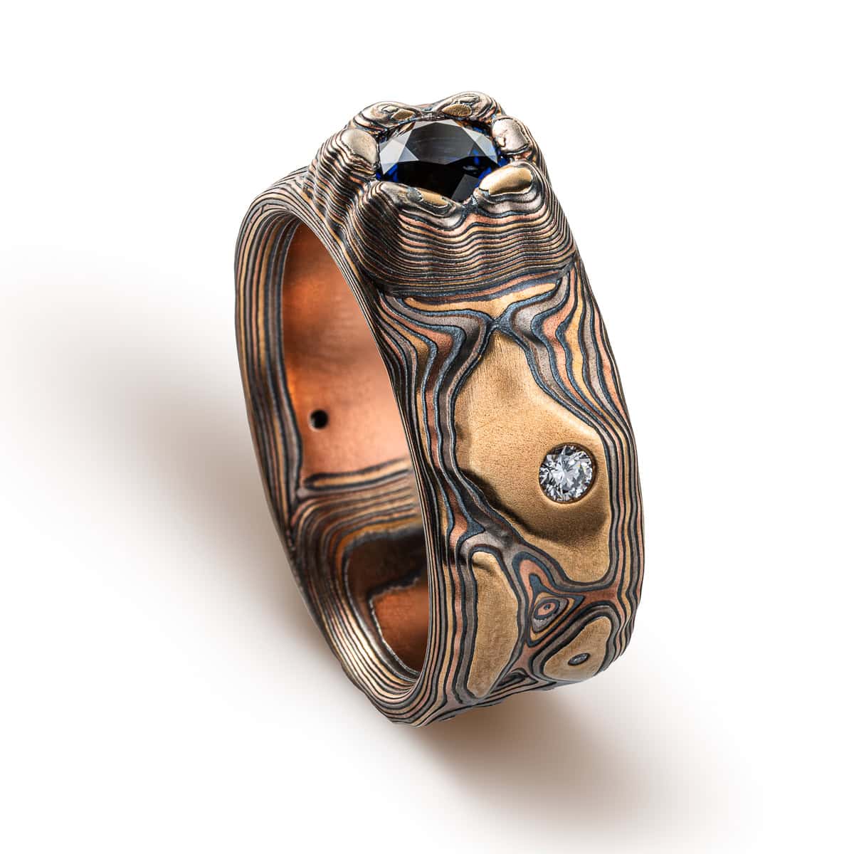 Large men's style mokume gane patterned ring, made to resemble rock formations and mountains, with a large dark blue and orange stone set into the ring in a raised area, intended to look like it's being excavated from the earth.