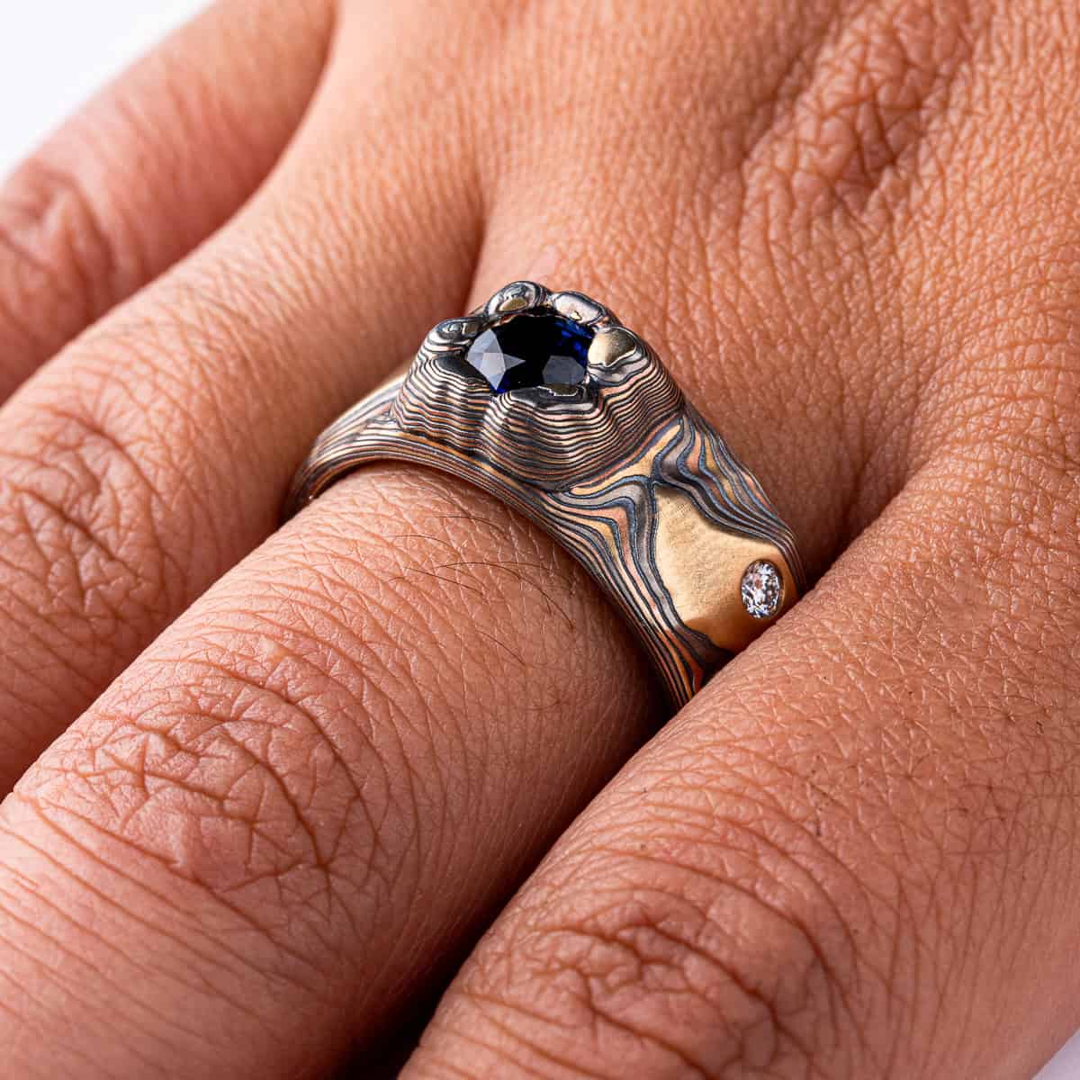 Large men's style mokume gane patterned ring, made to resemble rock formations and mountains, with a large dark blue and orange stone set into the ring in a raised area, intended to look like it's being excavated from the earth.