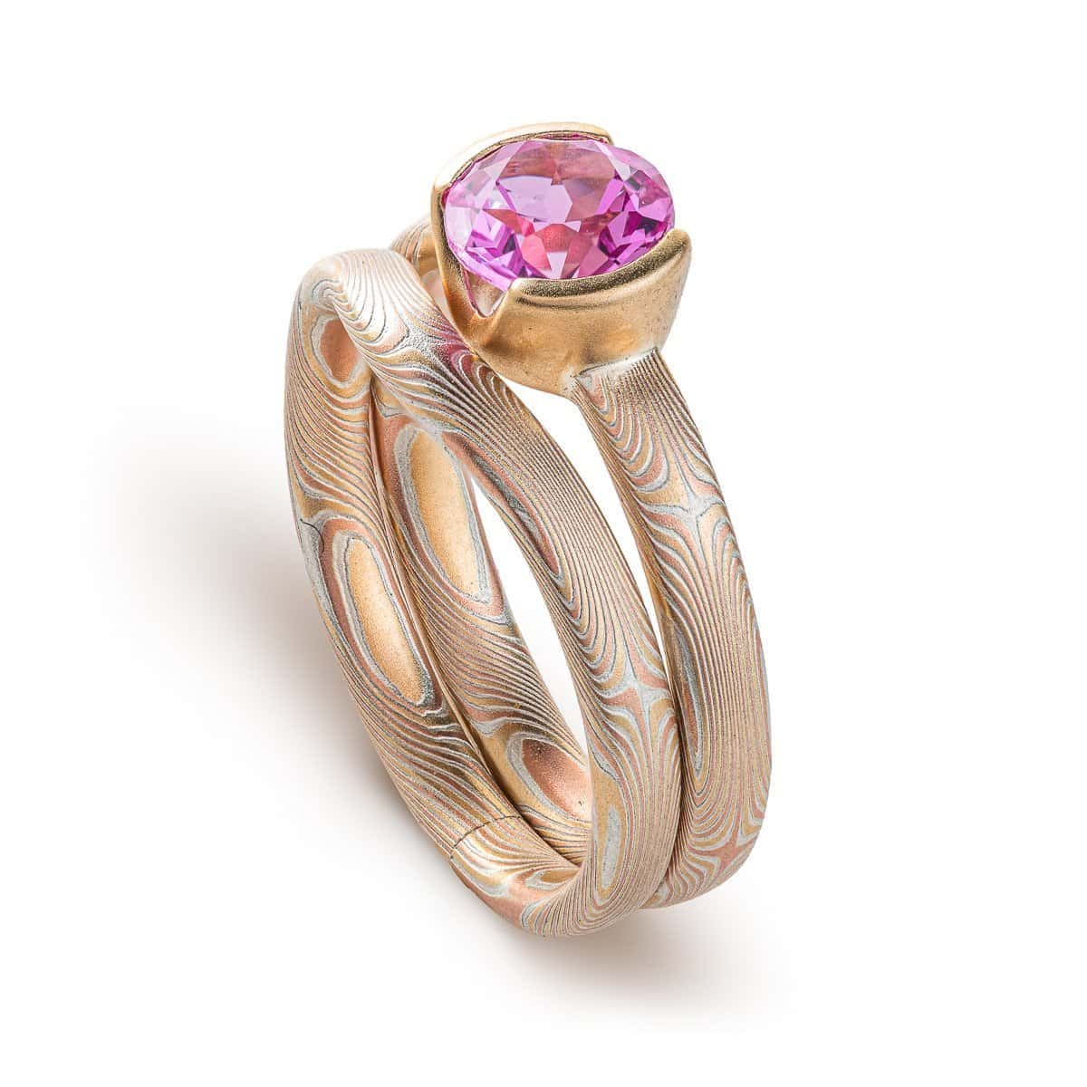 Pair of mokume gane patterned rings, one with a round pink stone, the other a band that curves around the stone and setting so the two can sit flush together. Overall warm light yellow gold look.