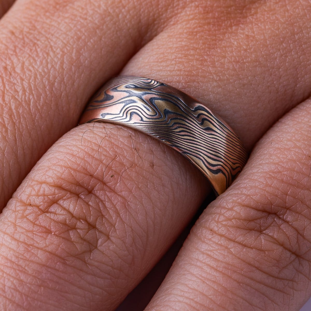 Wide mokume gane patterned ring, made with red and yellow gold and oxidized silver, with added carving to create a rippling, textured effect.