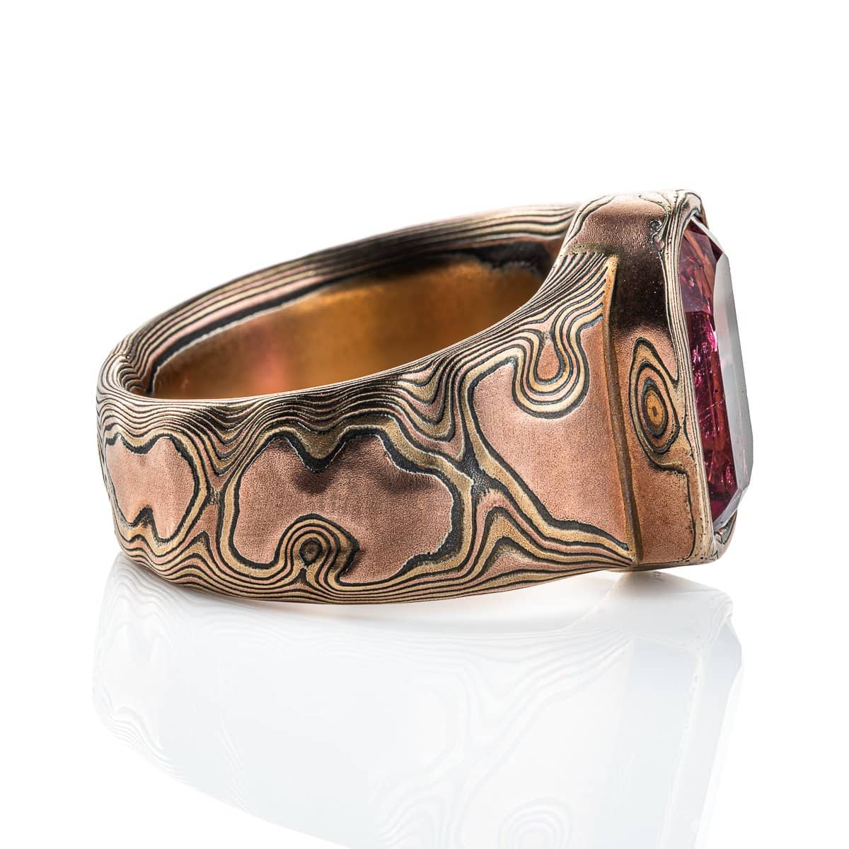 Mens mokume gane ring, with large pink stone surrounded by a border of mokume gane. The ring is wider at the top by the stone and tapers down towards the underside. Patterned in the woodgrain style, made with red gold, yellow gold and oxidized silver.