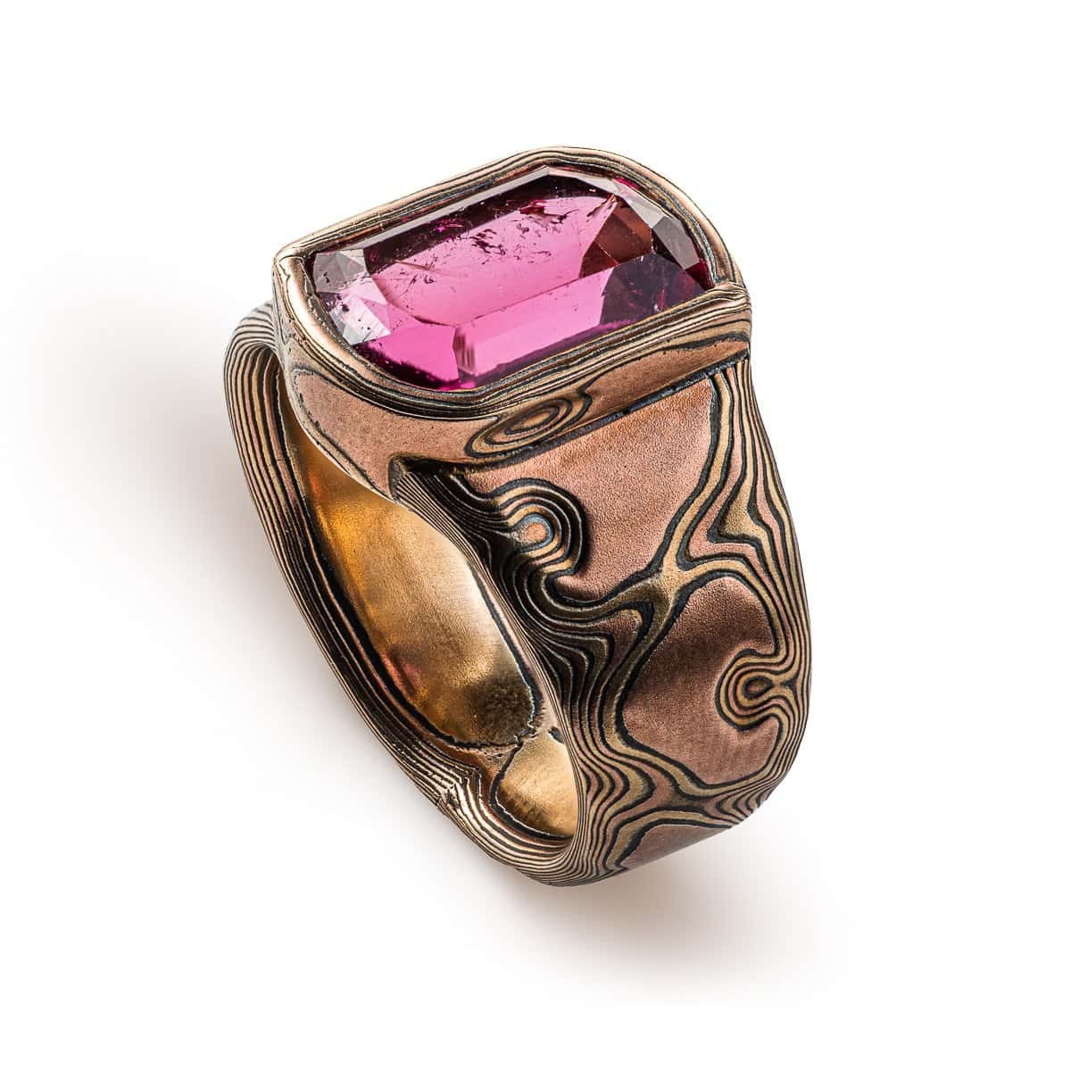 Mens mokume gane ring, with large pink stone surrounded by a border of mokume gane. The ring is wider at the top by the stone and tapers down towards the underside. Patterned in the woodgrain style, made with red gold, yellow gold and oxidized silver.