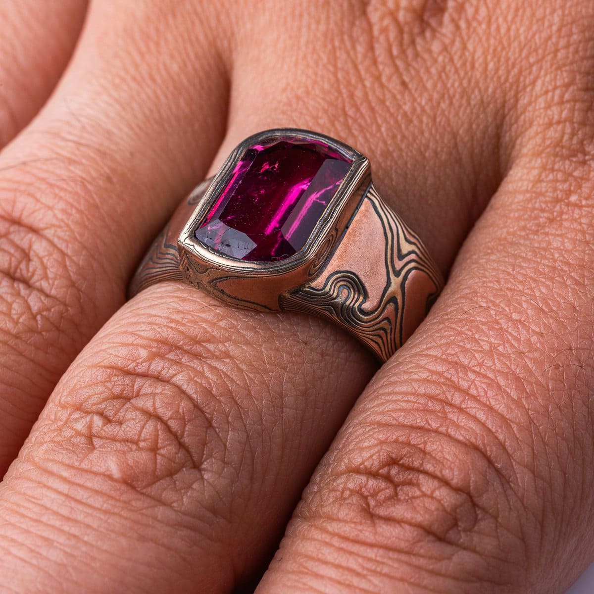 Mens mokume gane ring, with large pink stone surrounded by a border of mokume gane. The ring is wider at the top by the stone and tapers down towards the underside. Patterned in the woodgrain style, made with red gold, yellow gold and oxidized silver.