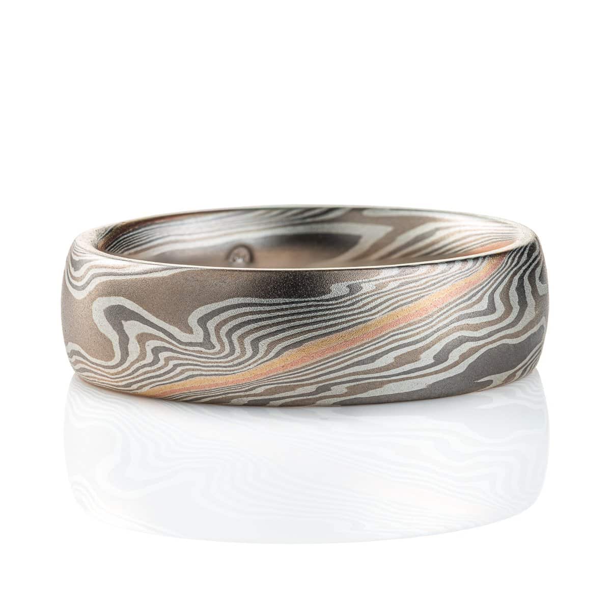Domed mokume gane band, with a smooth surface, the ring is mainly layers of dark and light gray, with a stripe of red gold and yellow gold at the center. The lines of the pattern run diagonally across and twisting around the ring.