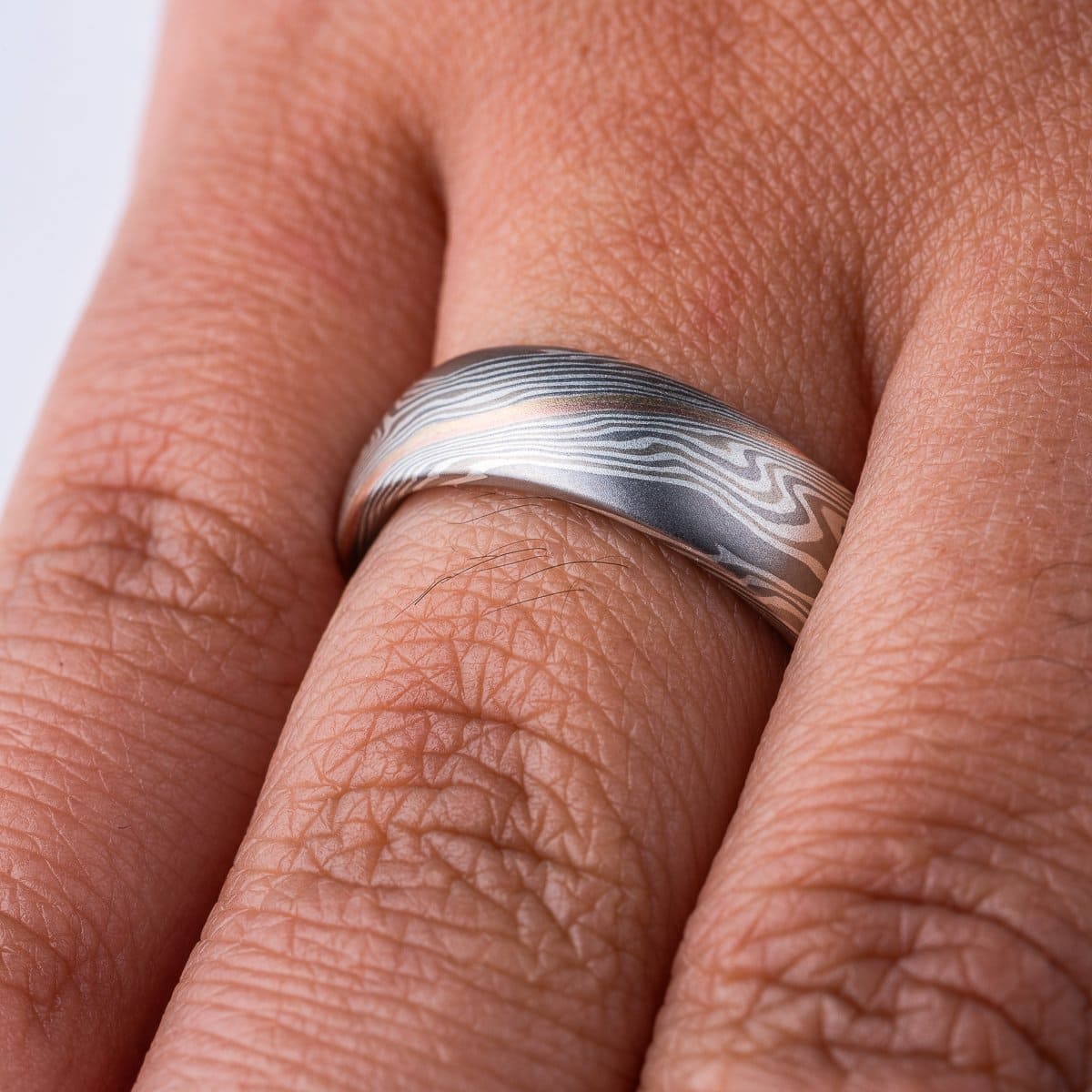 Domed mokume gane band, with a smooth surface, the ring is mainly layers of dark and light gray, with a stripe of red gold and yellow gold at the center. The lines of the pattern run diagonally across and twisting around the ring.