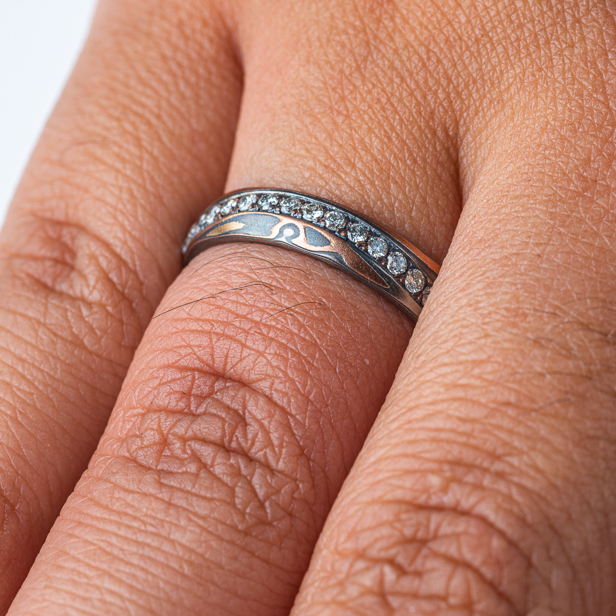Narrow mokume gane patterned band with undulating line of diamonds set flush into the surface. The band is made of warm toned metals in a woodgrain style pattern.